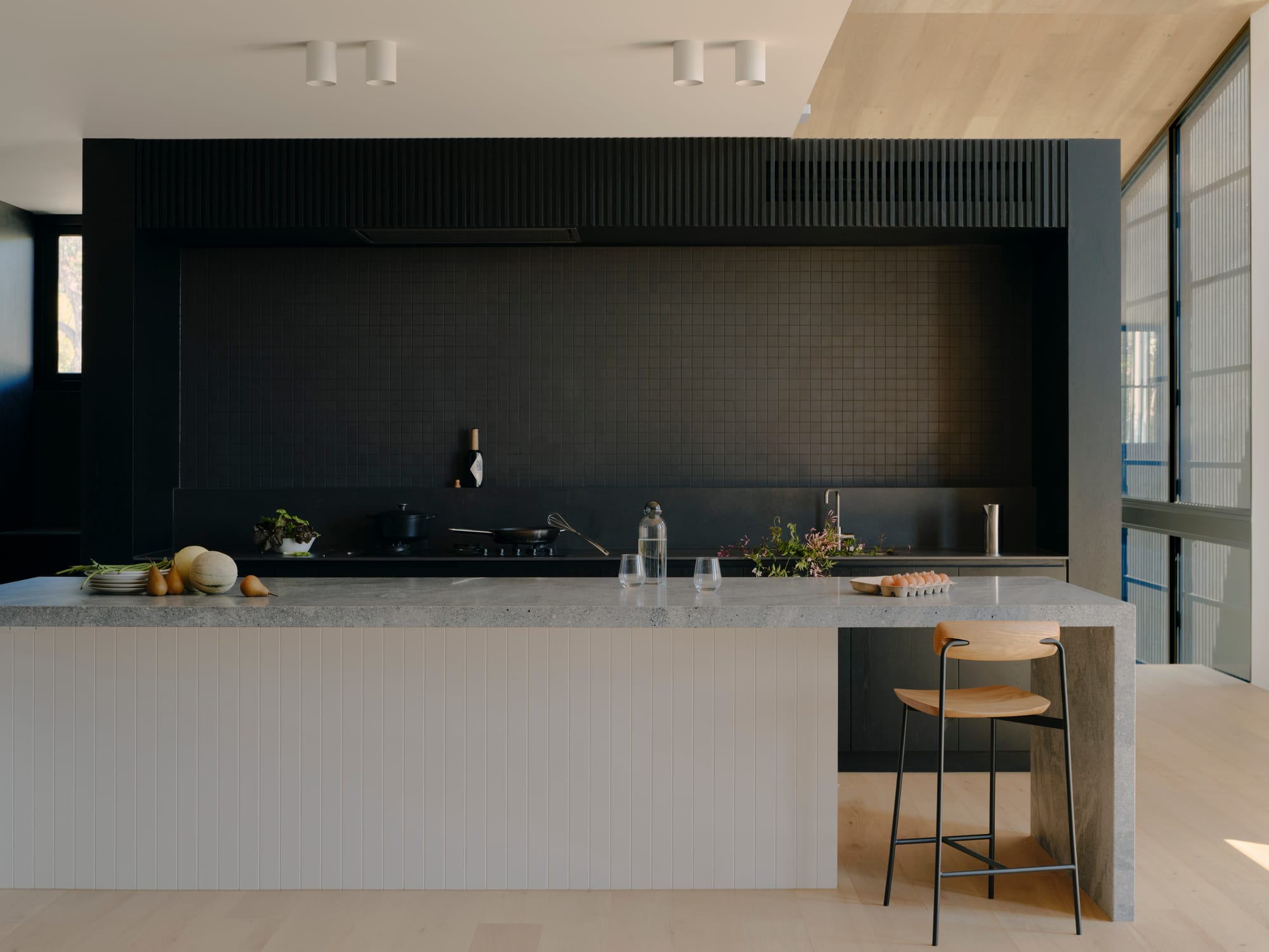 Courtside House by Tom Robertson Architects. Photography by Tom Ross. Modern residential kitchen with white timber clad island bench with granite benchtop. Black tiles splashback. Light timber floors.