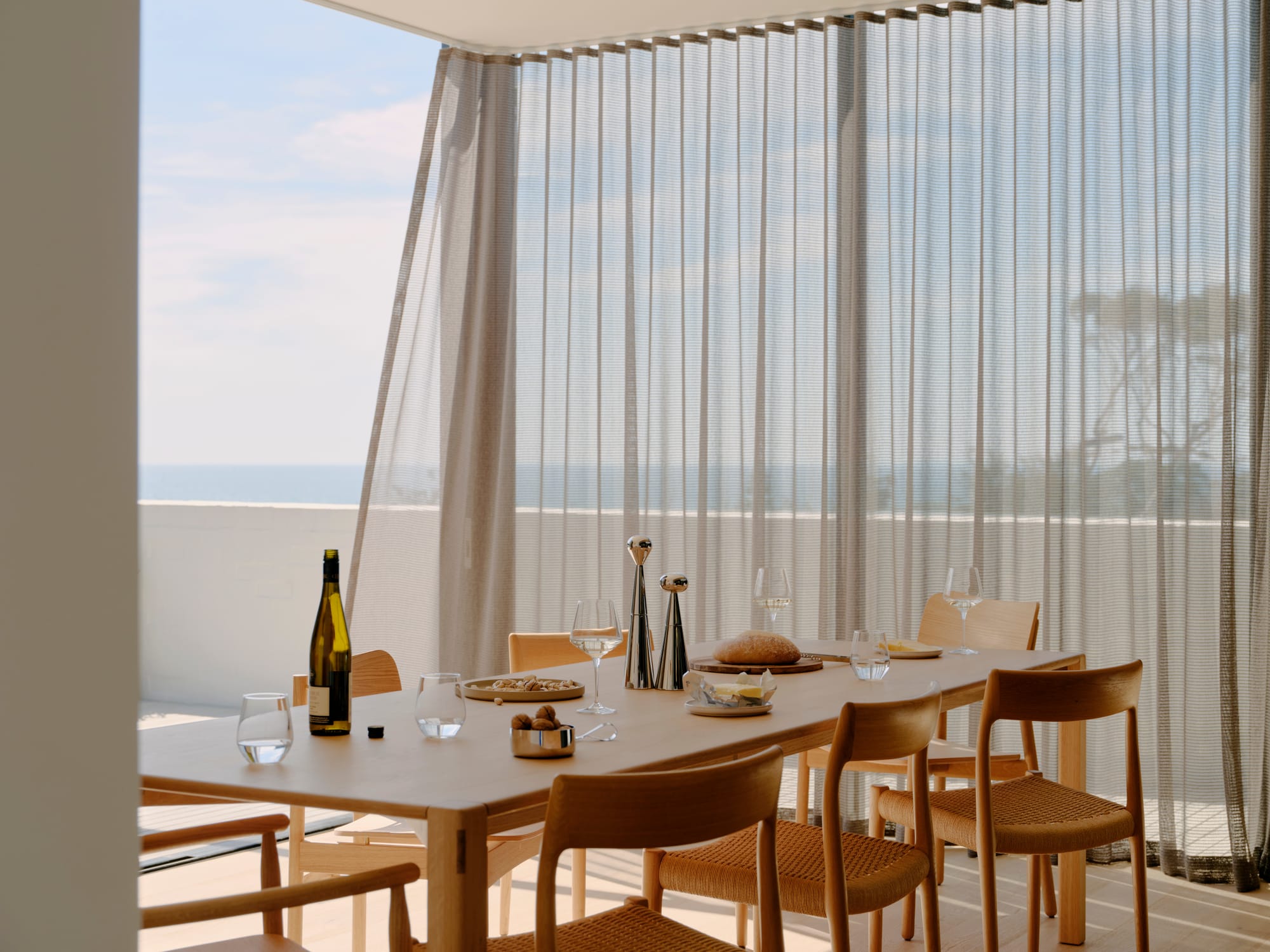 Courtside House by Tom Robertson Architects. Photography by Tom Ross. Dining room with floor-to-ceiling windows overlooking the ocean. Semi-transparent curtains blow in breeze behind minimalistic timber dining table and chairs. 