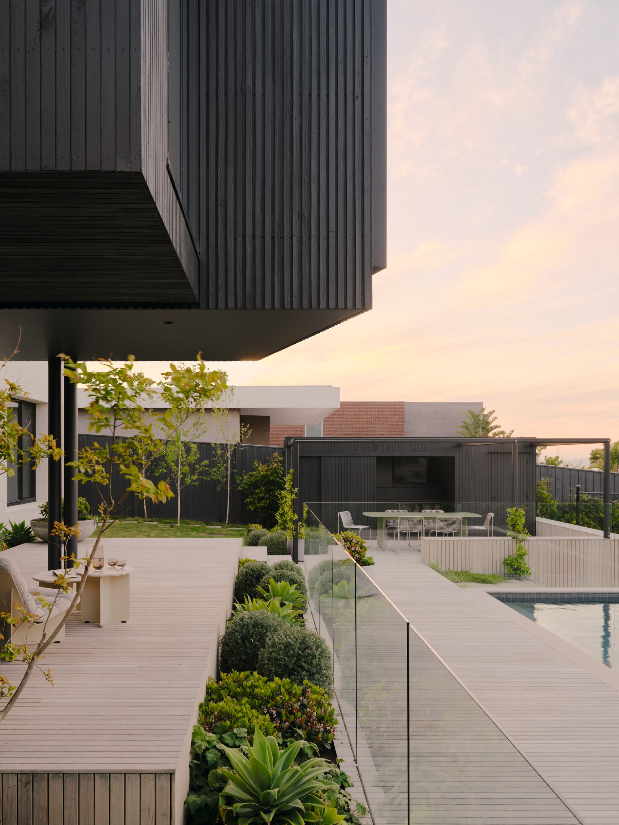 Courtside House by Tom Robertson Architects. Photography by Tom Ross. Exterior facade and gardens in elevation. Tiered decking leading to pool on right of image. Black pool house in right background. Grass and trees in left background if image. 