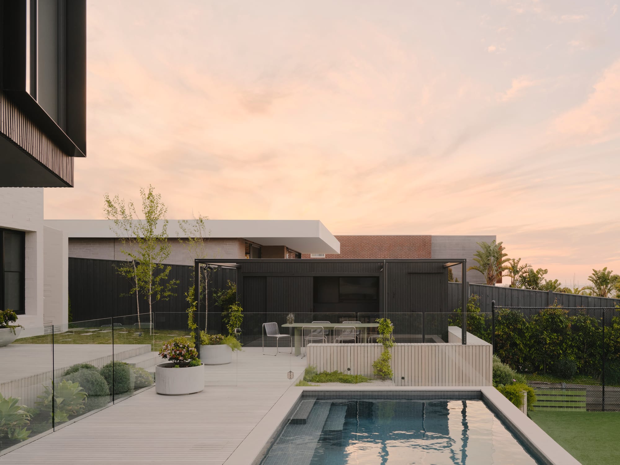 Courtside House by Tom Robertson Architects. Photography by Tom Ross. Backyard with light timber tiered decking leading to pool in centre of image. Lowered grass area to right of image and in left background. 