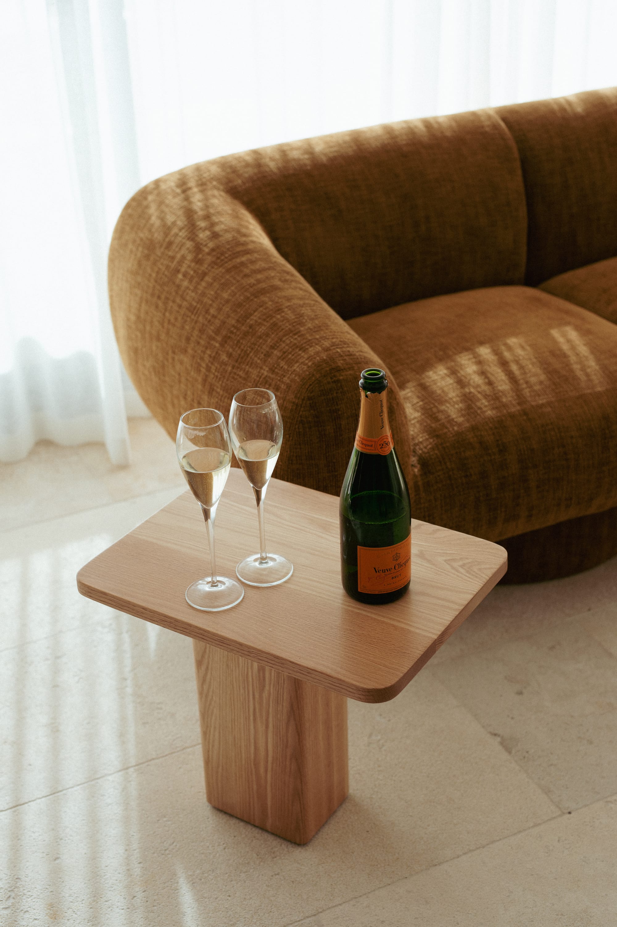 Made Furniture. Copyright of Made Furniture. Timber stool on tile floor with bottle of champagne and two glasses on it. Burnt orange sofa in background. 