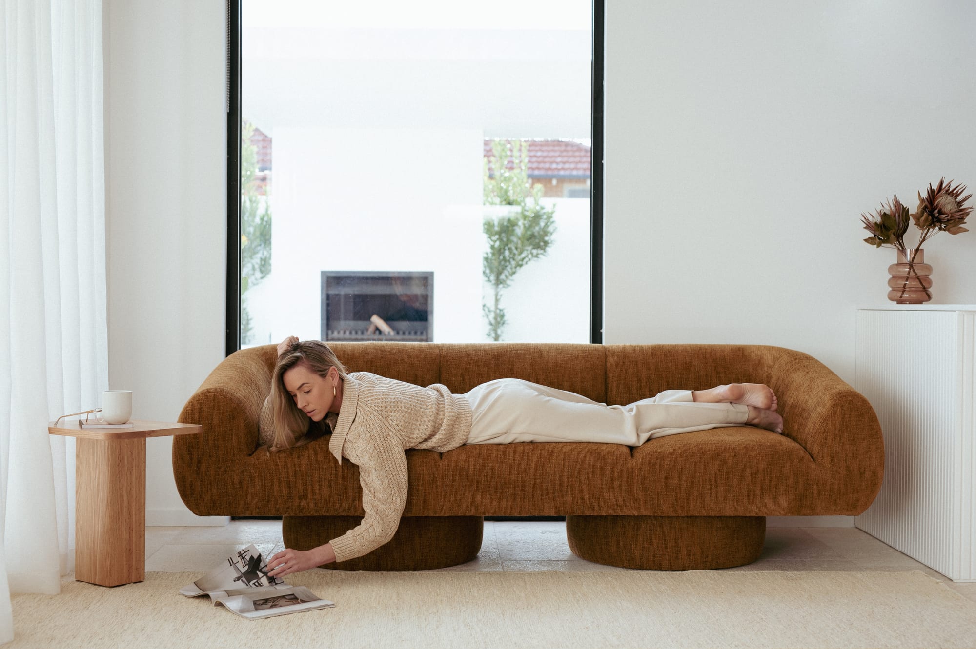A model laying on a Made Furniture sofa in a burnt orange fabric with a timber side table to the left and a large picture window in the background