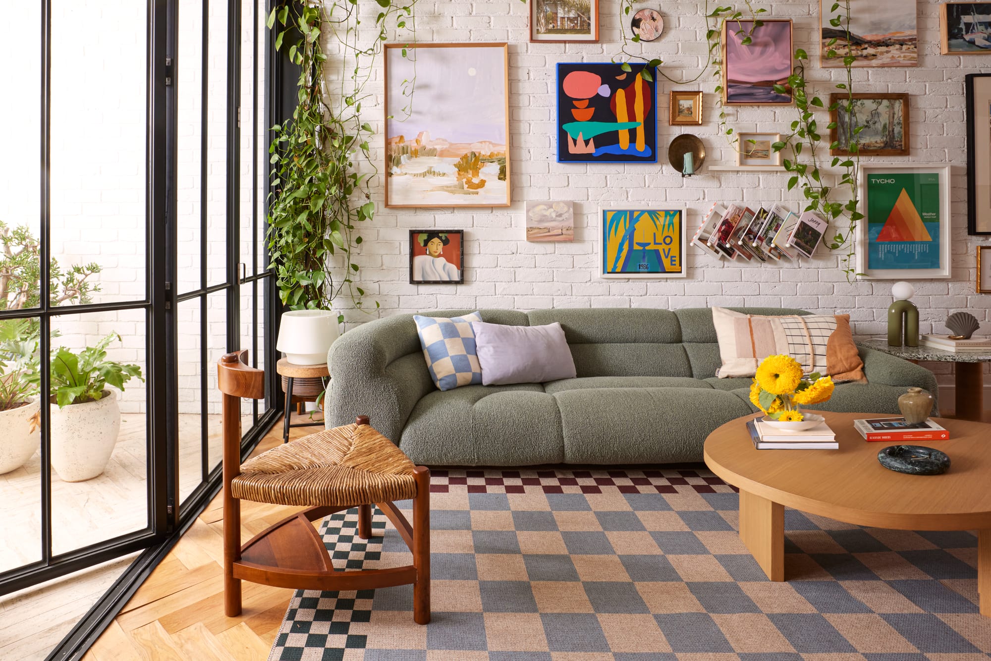 Double Rugs New Check Collection. Photographer: Jacqui Turk. Living room with green couch, white brick walls and herringbone timber floors. Checkered rug on floor. Lots of colourful artwork on wall. 