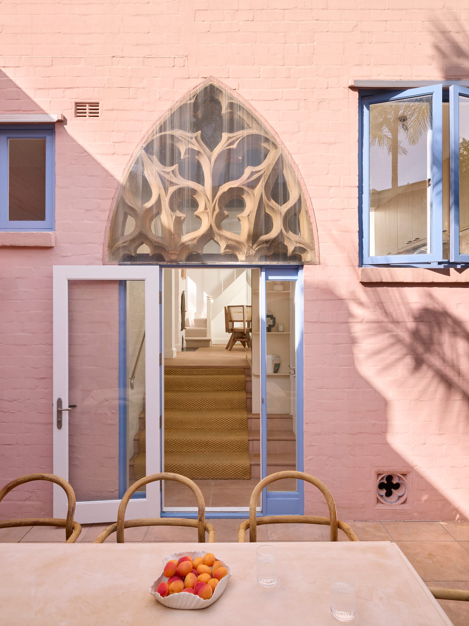 Chapel House by FURNISHD. Photography by Alicia Taylor. Gothic archway connecting inside and outside. Pink exterior brick walls with blue window and door frames. Dining table with cane chairs in foreground.  