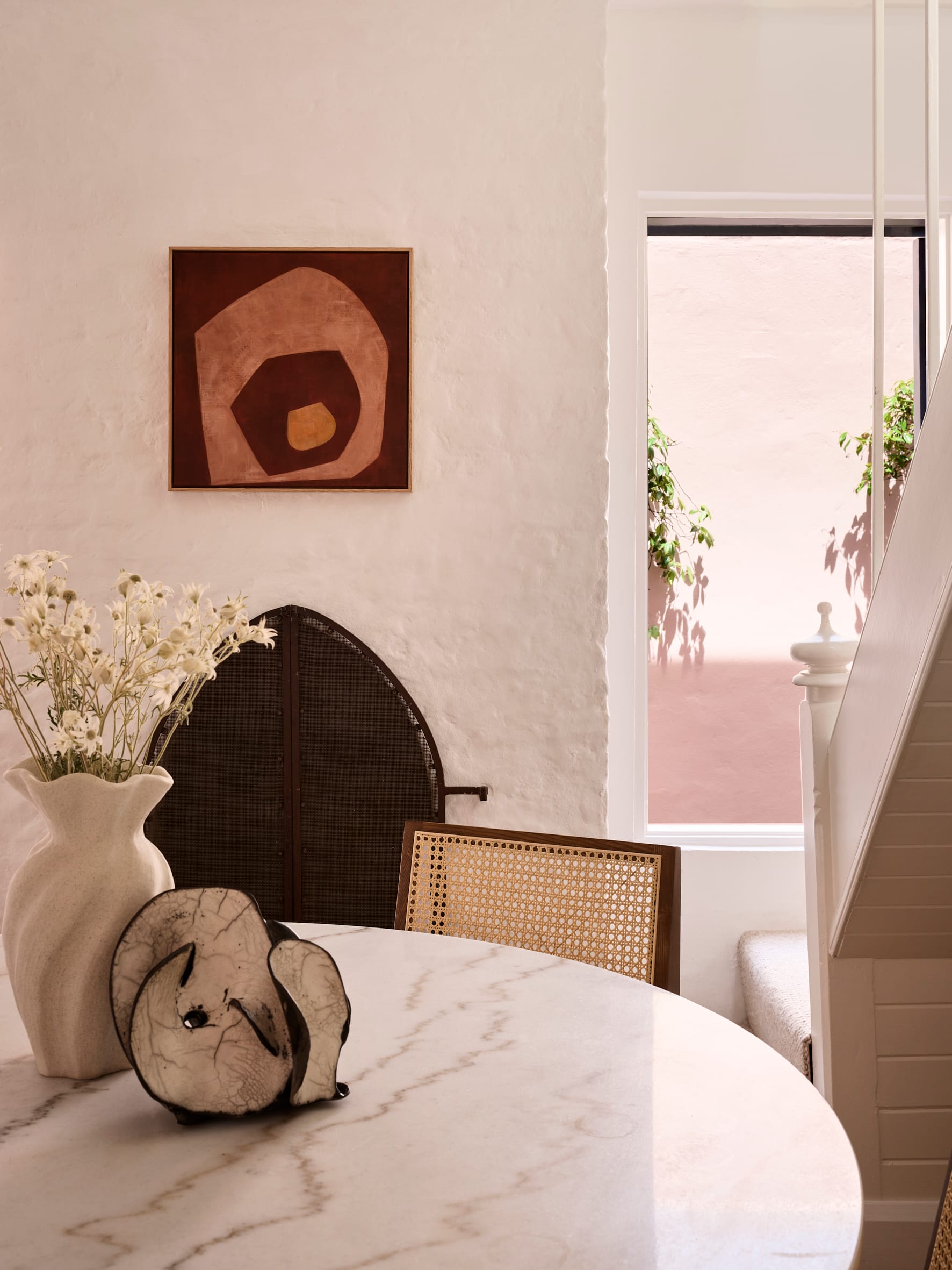 Chapel House by FURNISHD. Photography by Alicia Taylor. Round marble table with rattan chair. White brick wall in background with window looking onto pink outside wall.