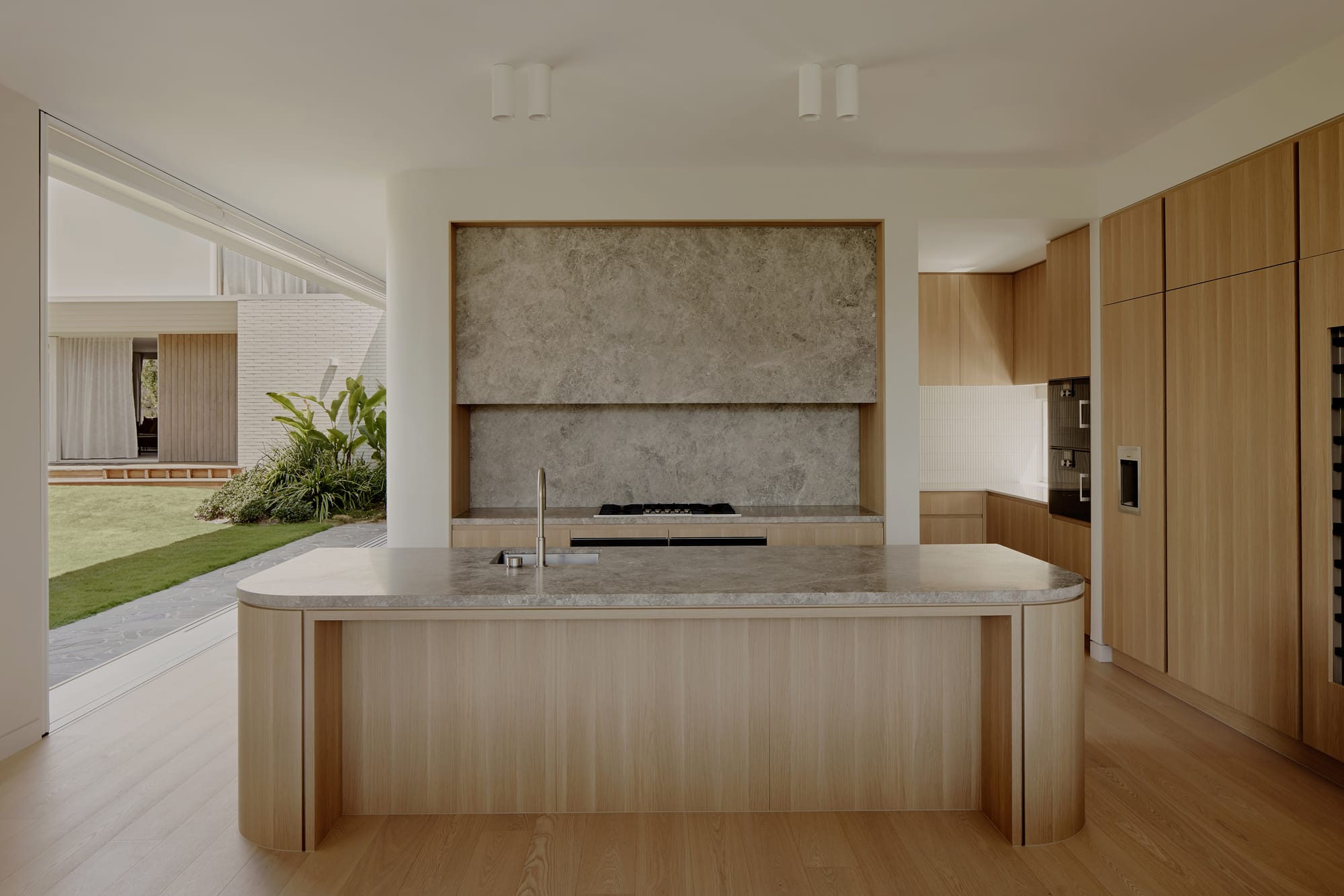 Hill Courtyard House by Kelder Architects. Photography by Brock Beazley Photography. Kitchen with timber floors, island bench, joinery. Stone benchtop and splashback. Opening onto green courtyard to the left. 