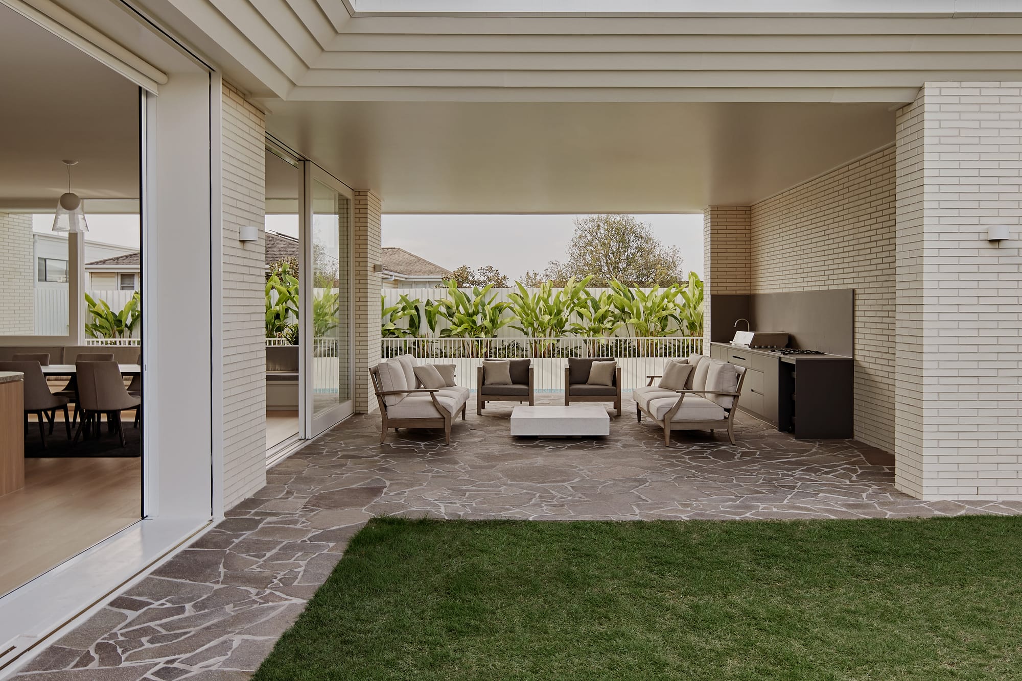 Hill Courtyard House by Kelder Architects. Photography by Brock Beazley Photography. Undercover patio with white brick walls, white-framed sliding doors and stone pavers. White outdoor furniture arranged around white coffee table. 