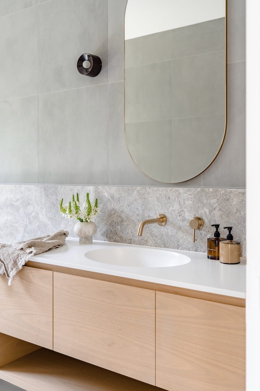 Kasa Byron Bay. Photography by Adam Nicolson. Timber bathroom vanity with white countertop and integrated sink. Stone splashback and grey tiled wall.  Large gold framed round mirror.