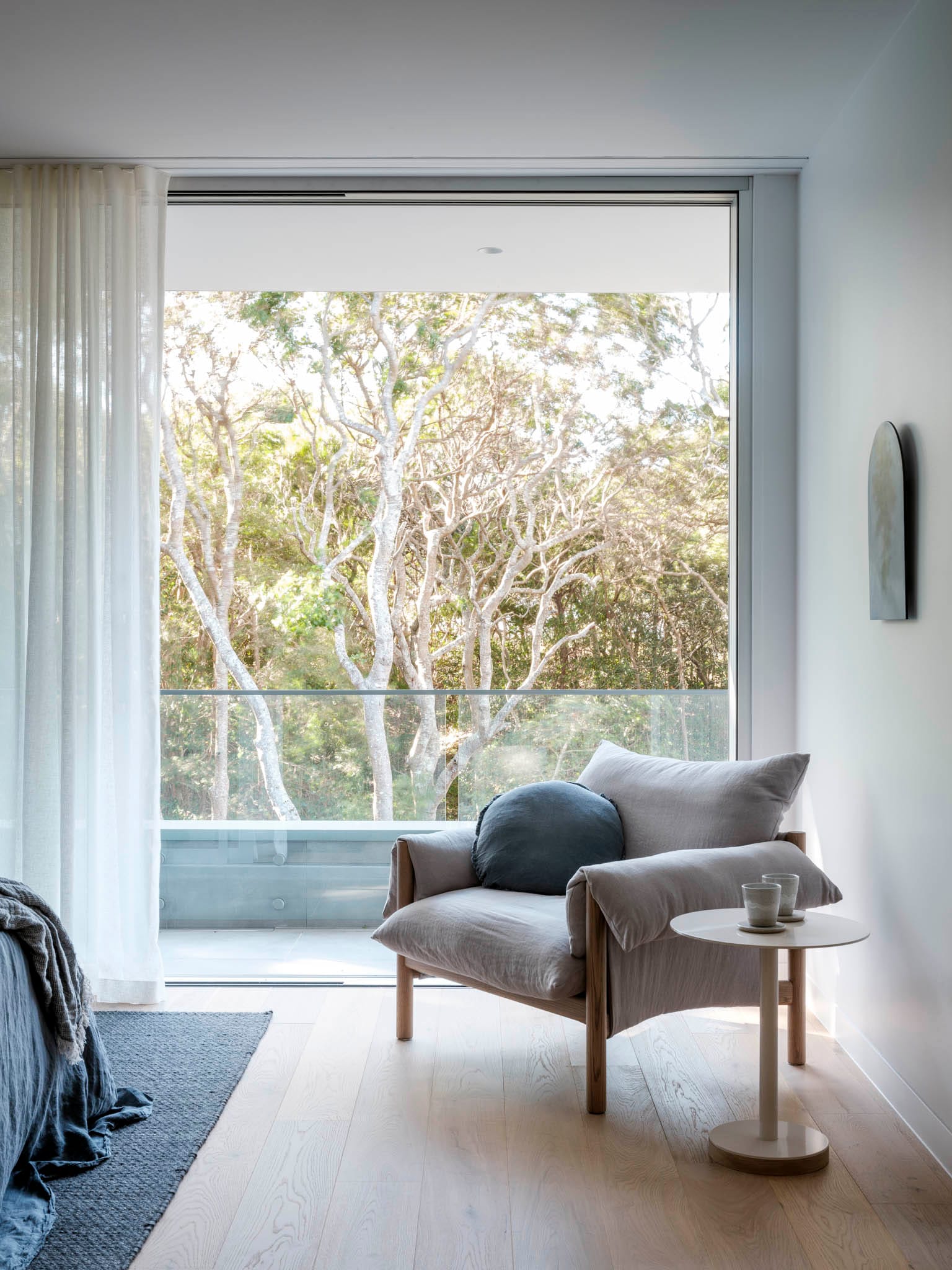 Kasa Byron Bay. Photography by Tom Ferguson. Blush and timber armchair with side table in front of floor-to-ceiling window overlooking trees. Timber floors in room.