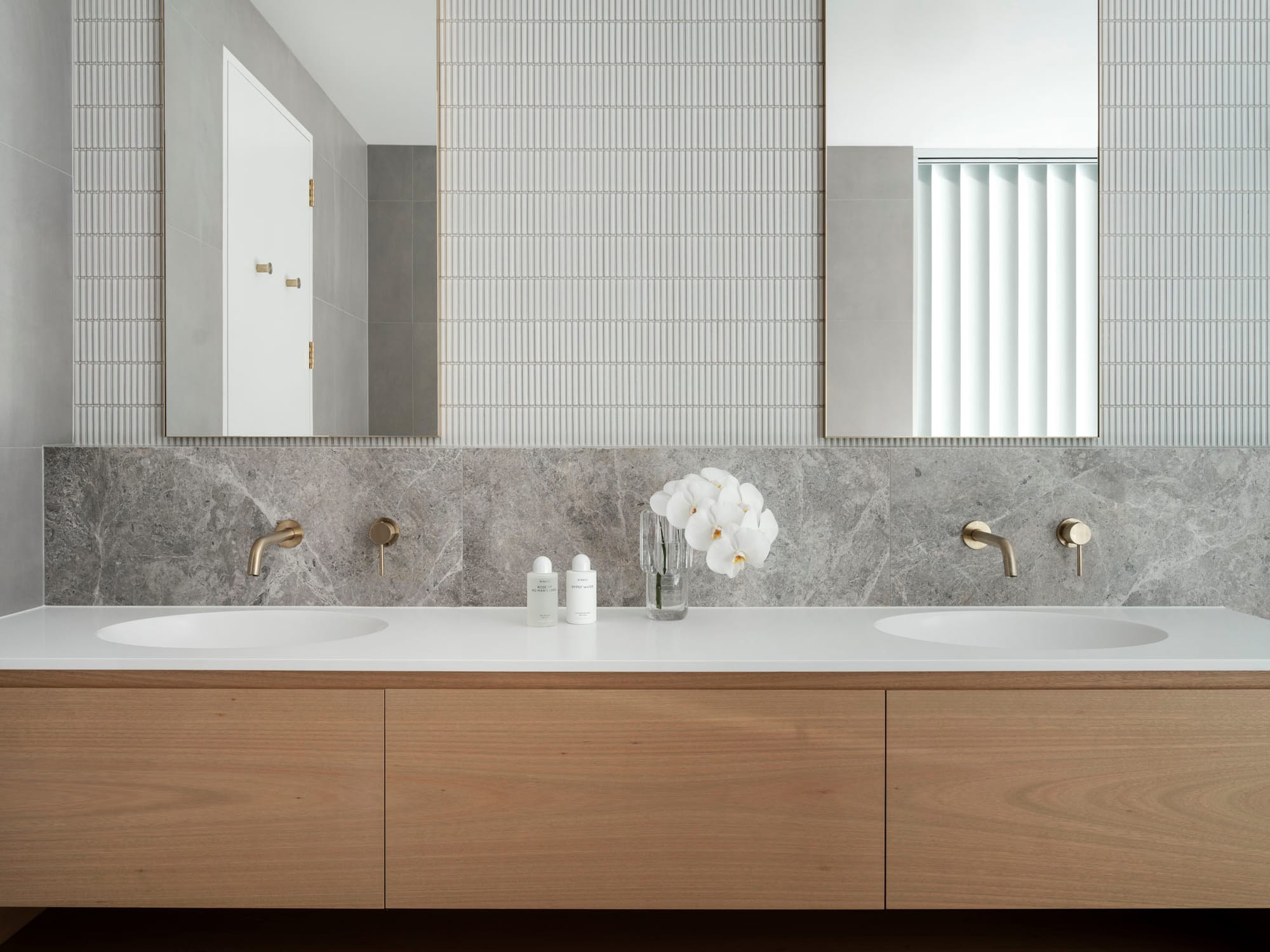 Kasa Byron Bay. Photography by Tom Ferguson. Timber bathroom vanity with stone splashback and tiled wall. Double gold framed mirrors above double white sinks. 