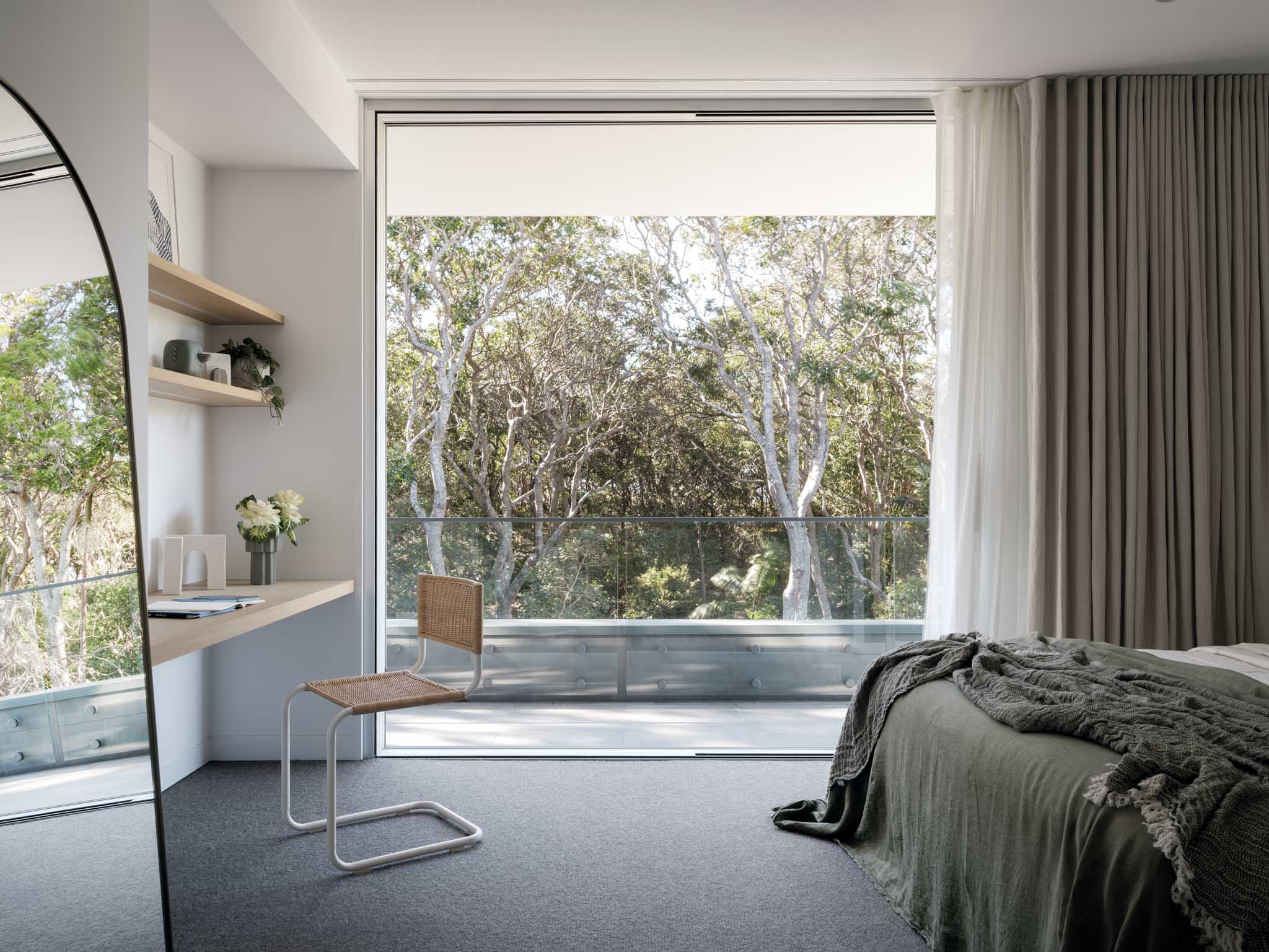 Kasa Byron Bay. Photography by Tom Ferguson. Bedroom overlooking treetops from adjoining balcony. Dark carpets and beige curtains. Study nook and mirror to left of image. 