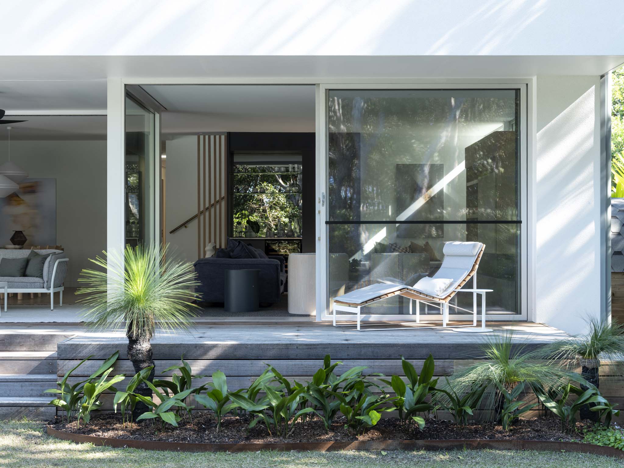 Kasa Byron Bay. Photography by Tom Ferguson. Timber patio opening up from interior living space.  White reclining deck chair. Garden bed below deck. 
