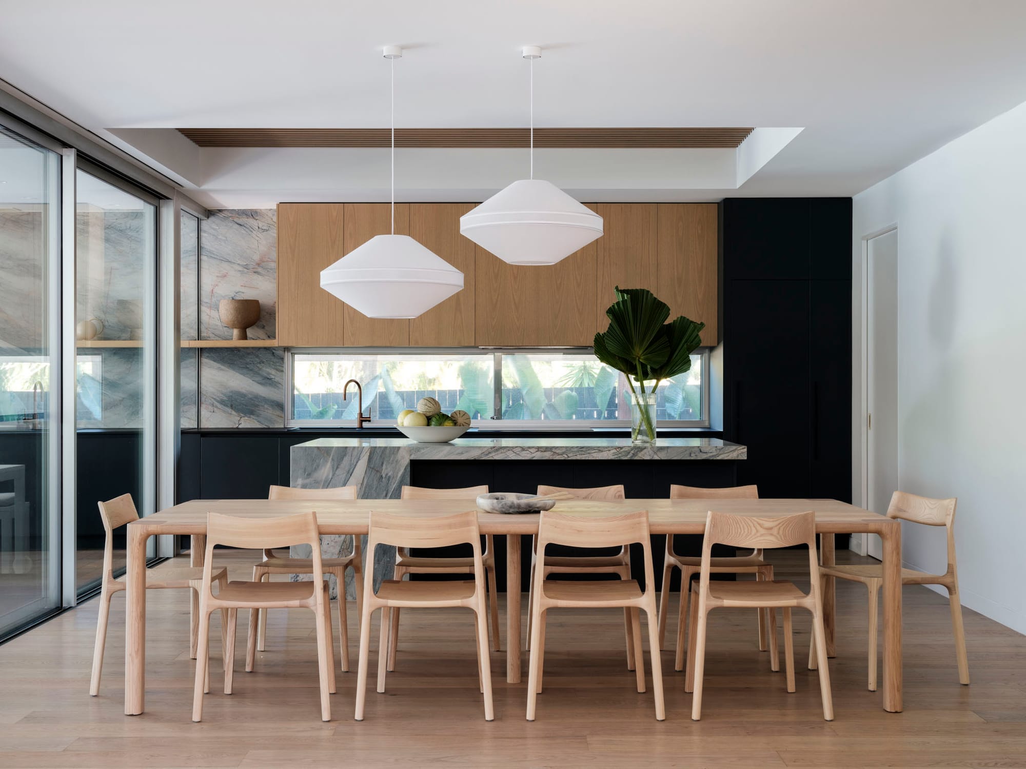Kasa Byron Bay. Photography by Tom Ferguson. Large timber dining setting in front of contemporary kitchen. Black cabinetry and stone splashback and countertops. Two large white pendant lights.