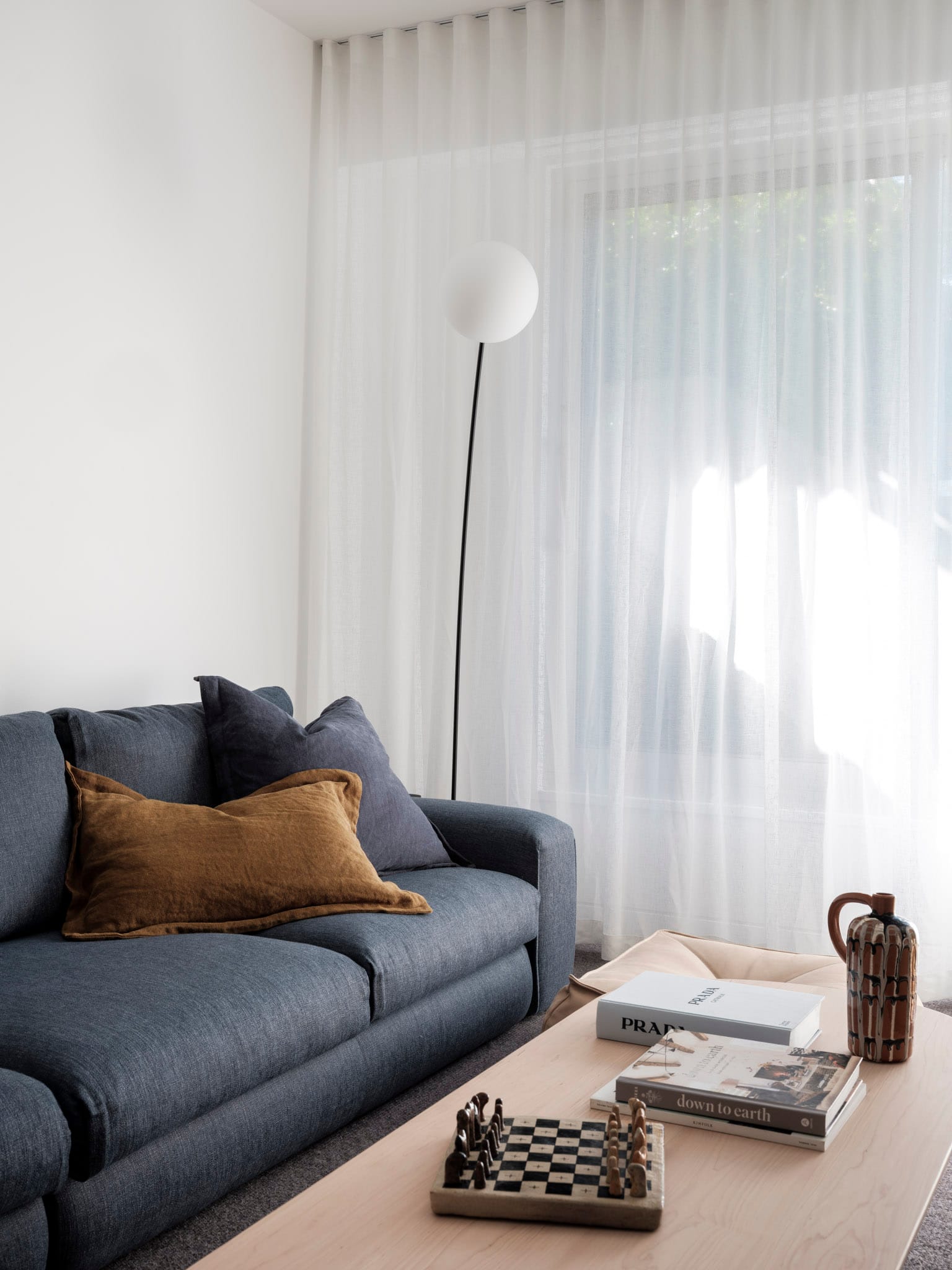 Kasa Byron Bay. Photography by Tom Ferguson. Navy couch and timber coffee table. White curtains in background. Simple lamp with black stem and white orb. 