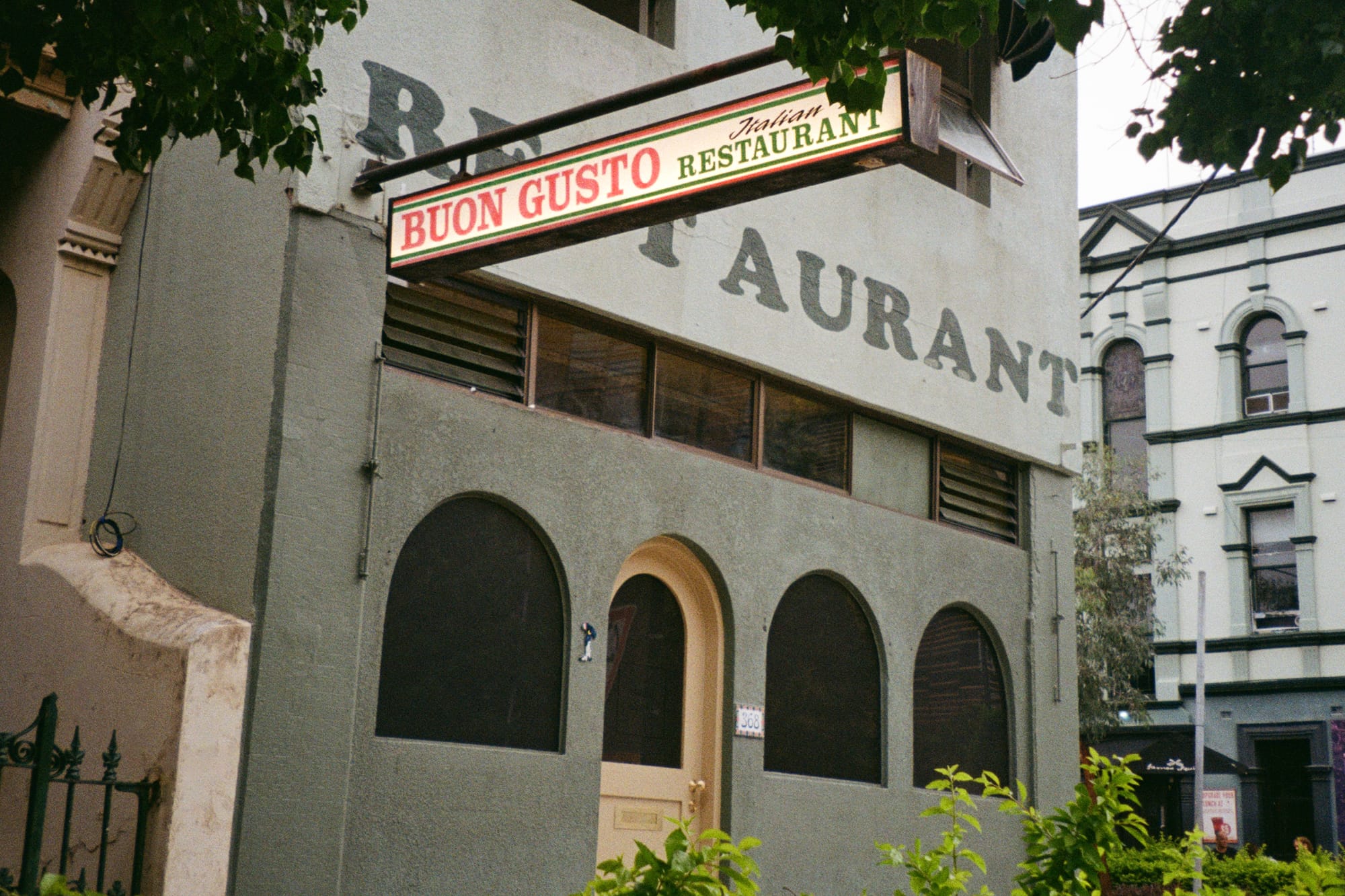 Buon Gusto. Photography by Benjamin Jay Shand. Facade of double storey building with arched door and windows. Italian restaurant sign hanging off wall. Dark green exterior.