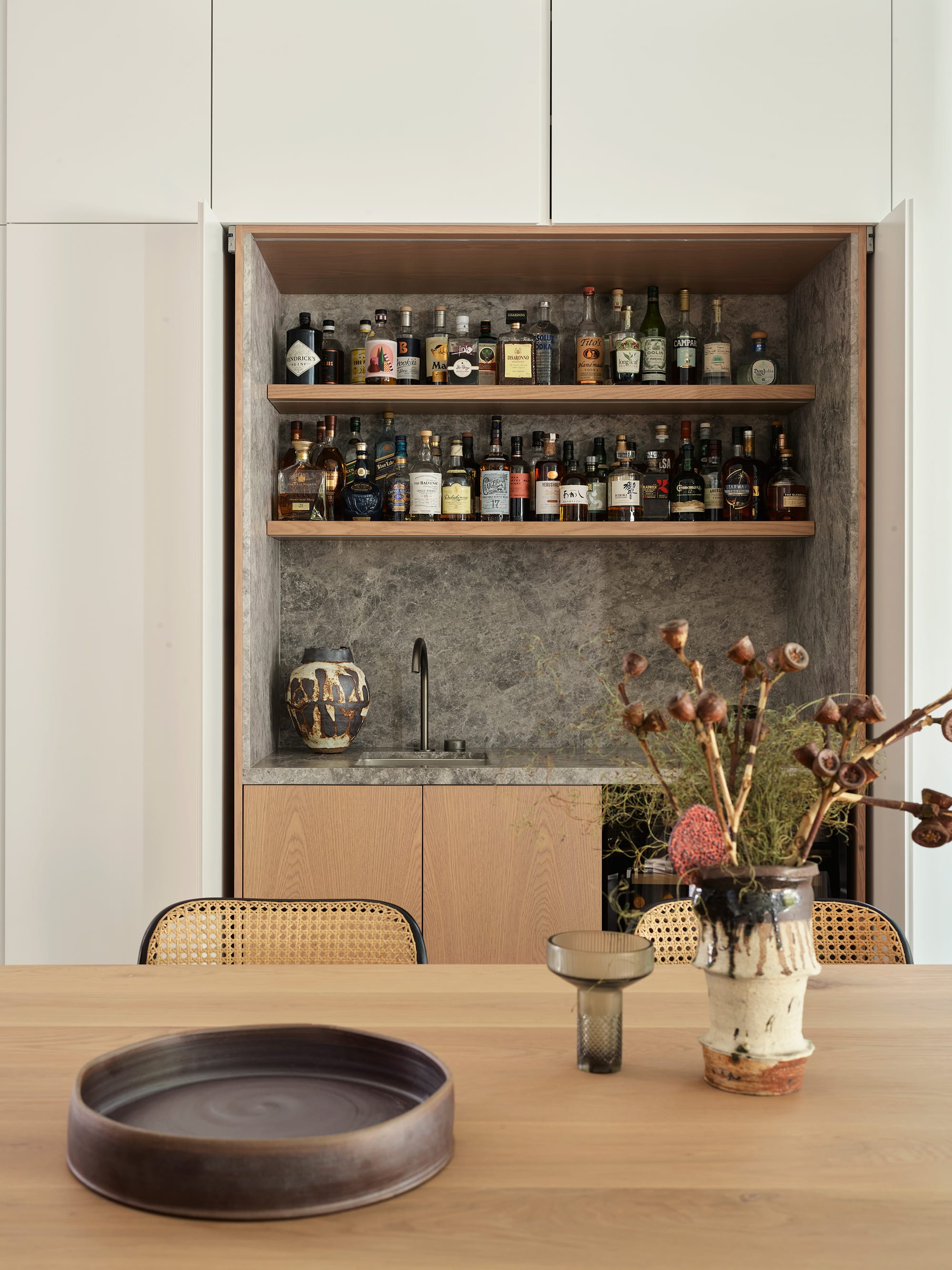 Warren House by CM Studio. Photography by Nic Gossage. Bar counter with grey countertop and splashback. Timber table in front with rattan chairs.
