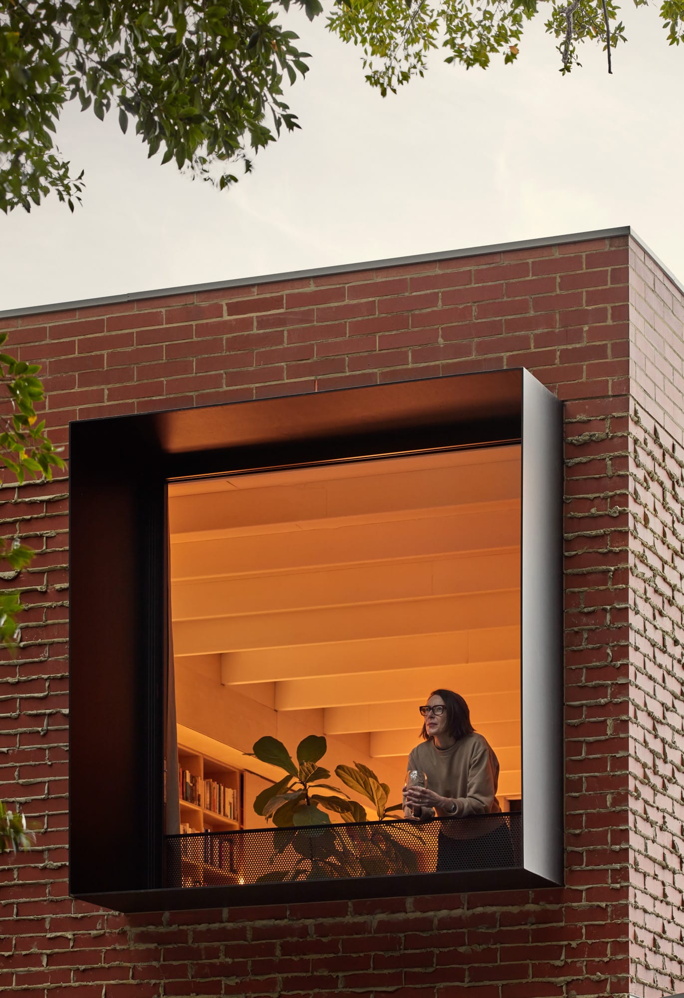 Brick House by Studio Roam. Photography by Jack Lovel. Double storey brick facade of home with large window boxes on ground and first floor. Warm lighting coming from black framed windows and doors. 
