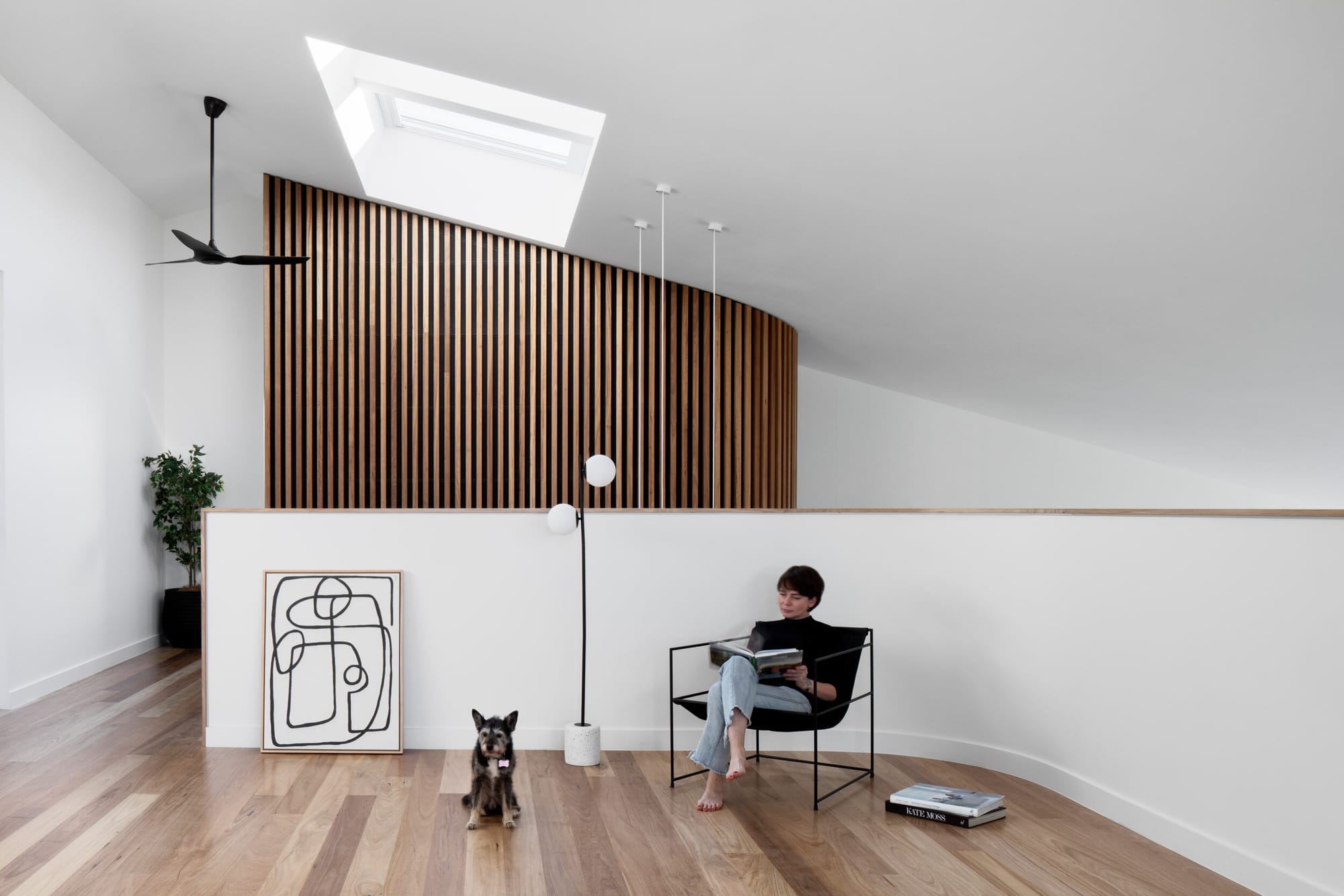 Dickens Street Residence by Chan Architecture. Photography by Tatjana Plitt. Woman sitting in armchair reading. White, curved half wall behind her seat with full-height timber clad wall in background. 