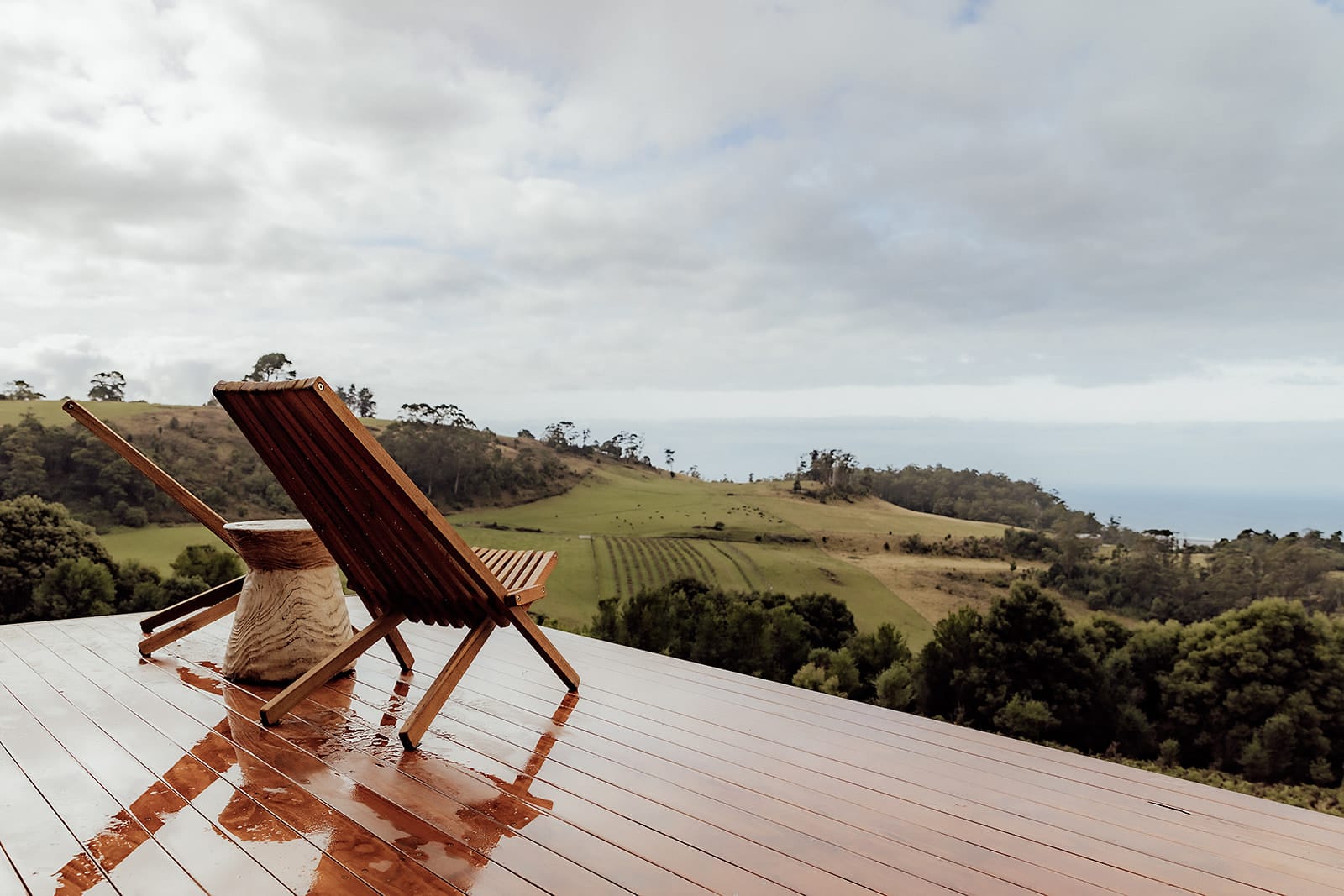 Candlebark Ridge. Copyright of Candlebark Ridge. Views of rilling grassy hills and ocean from timber deck. Two deck chairs and side table in foreground. 