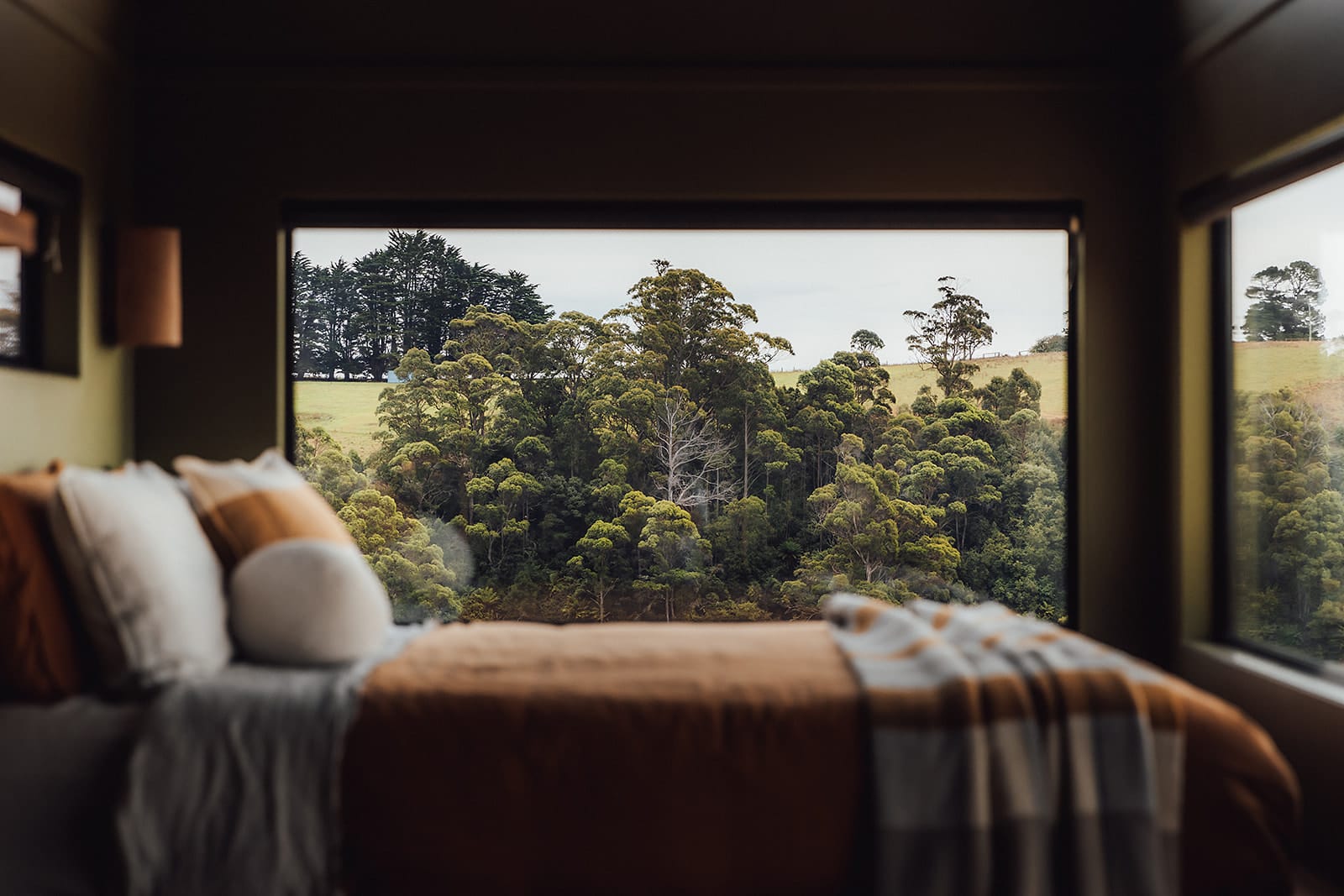 Candlebark Ridge. Copyright of Candlebark Ridge. Views of green hills and native bushland out of window. Bed with orange and blue bedding unfocused in foreground. 