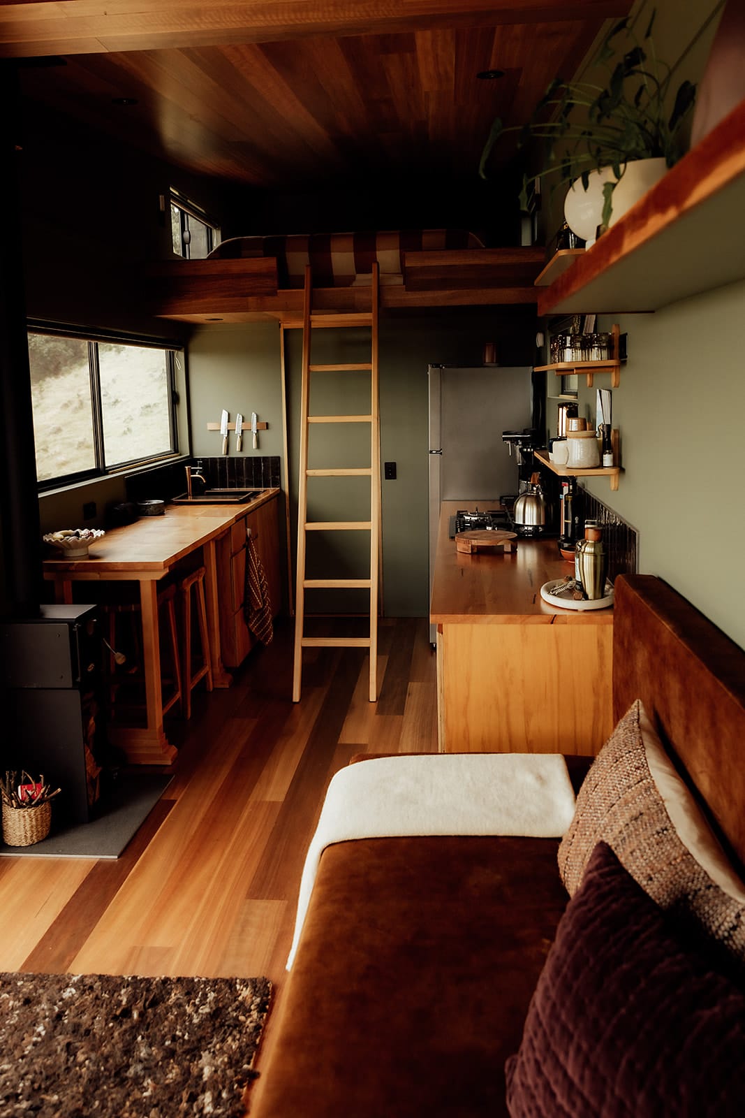 Candlebark Ridge. Copyright of Candlebark Ridge. Kitchen, dining and living area in tiny home. Black fireplace to left. Timber floors and joinery with green walls. 