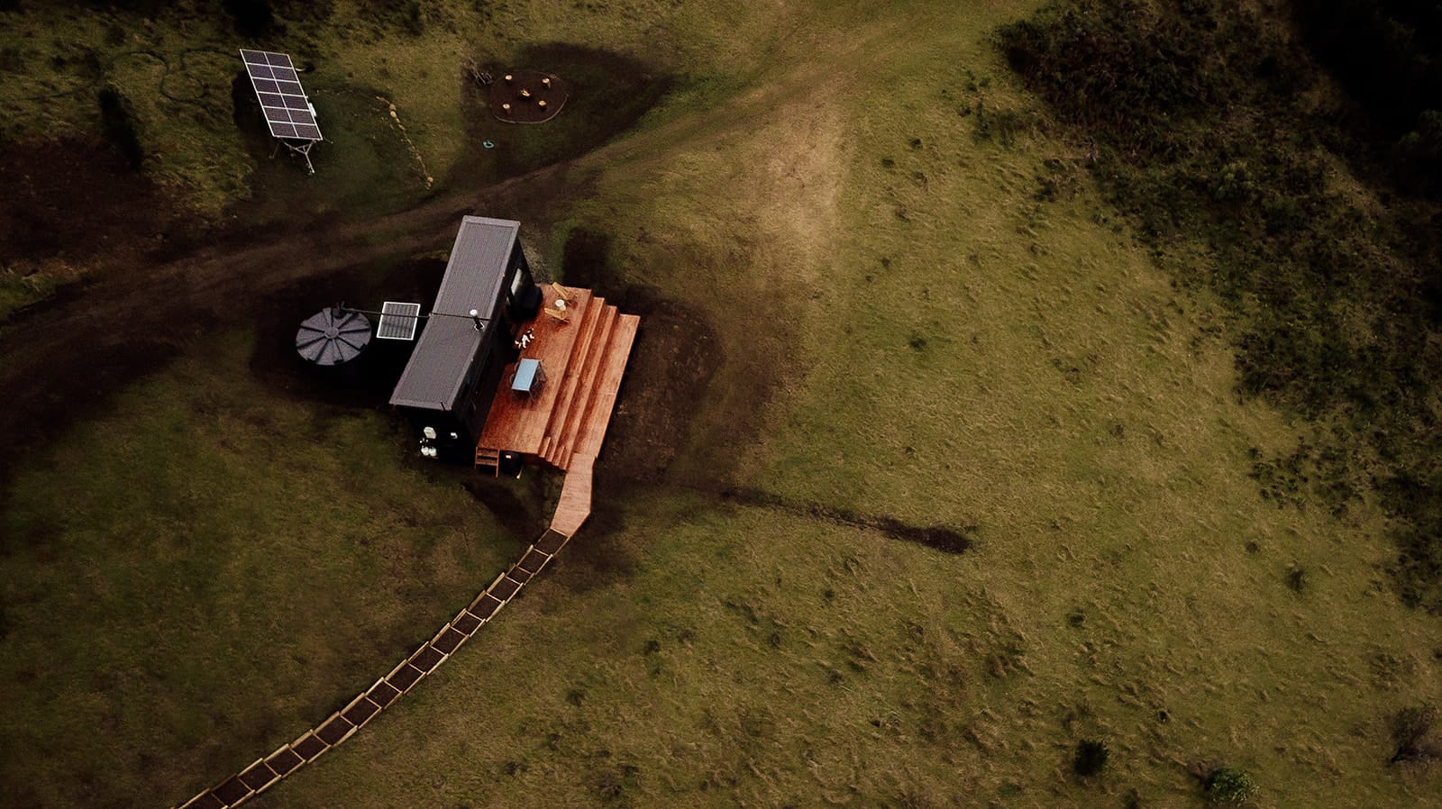 Candlebark Ridge. Copyright of Candlebark Ridge. Aerial shot of tiny home with large timber deck in rolling green paddock. 