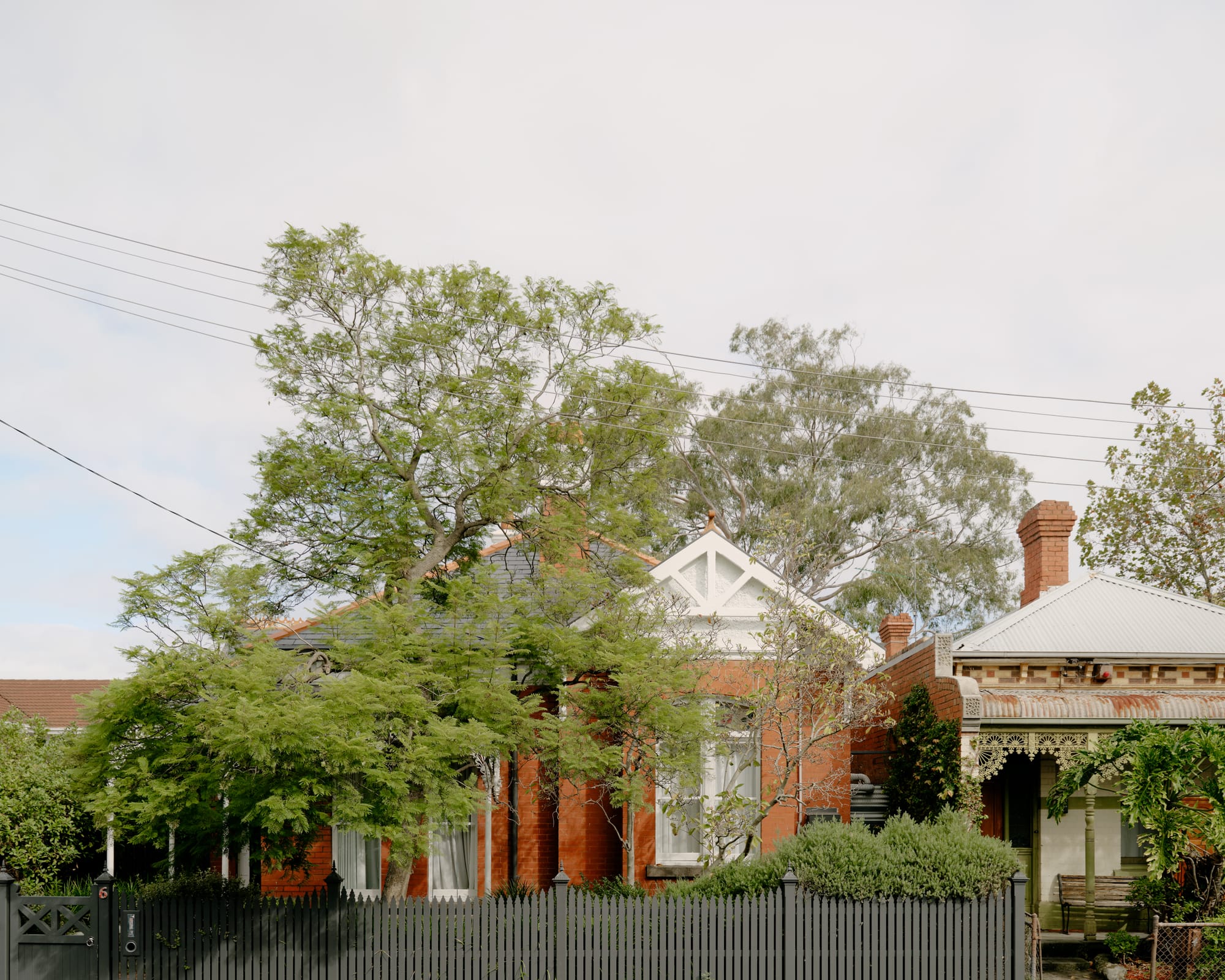 Corymbia by Karen Abernethy Architects. Photography by Tom Ross. Street view of Victorian-era red brick home with pitched roof and grey picket fence. Jacaranda tree growing out the front of home. 