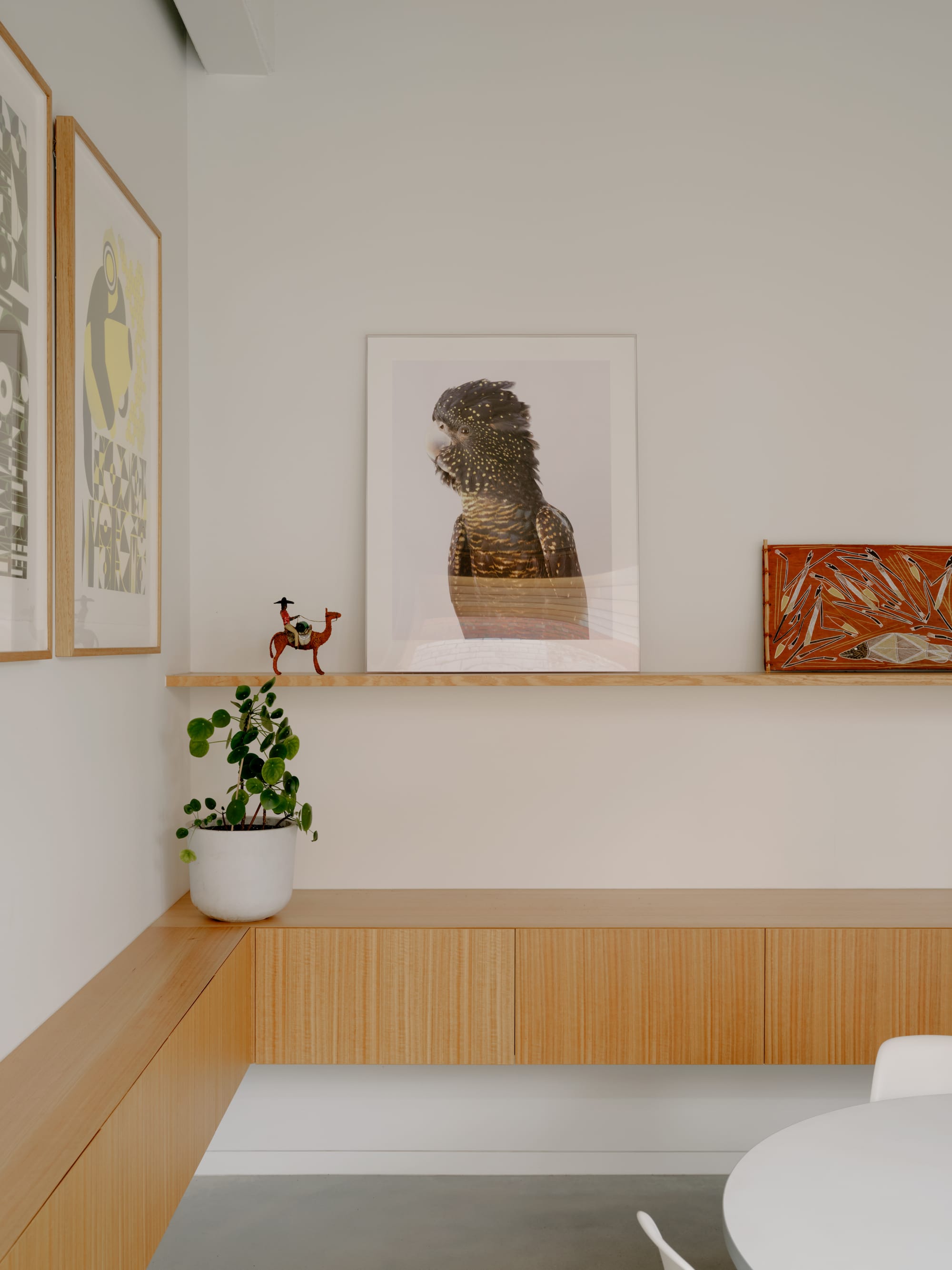 Corymbia by Karen Abernethy Architects. Photography by Tom Ross. Timber joinery wrapping around corner of two walls. Timber picture shelves display several eclectic images. White walls and white table and chairs visible in bottom right corner,