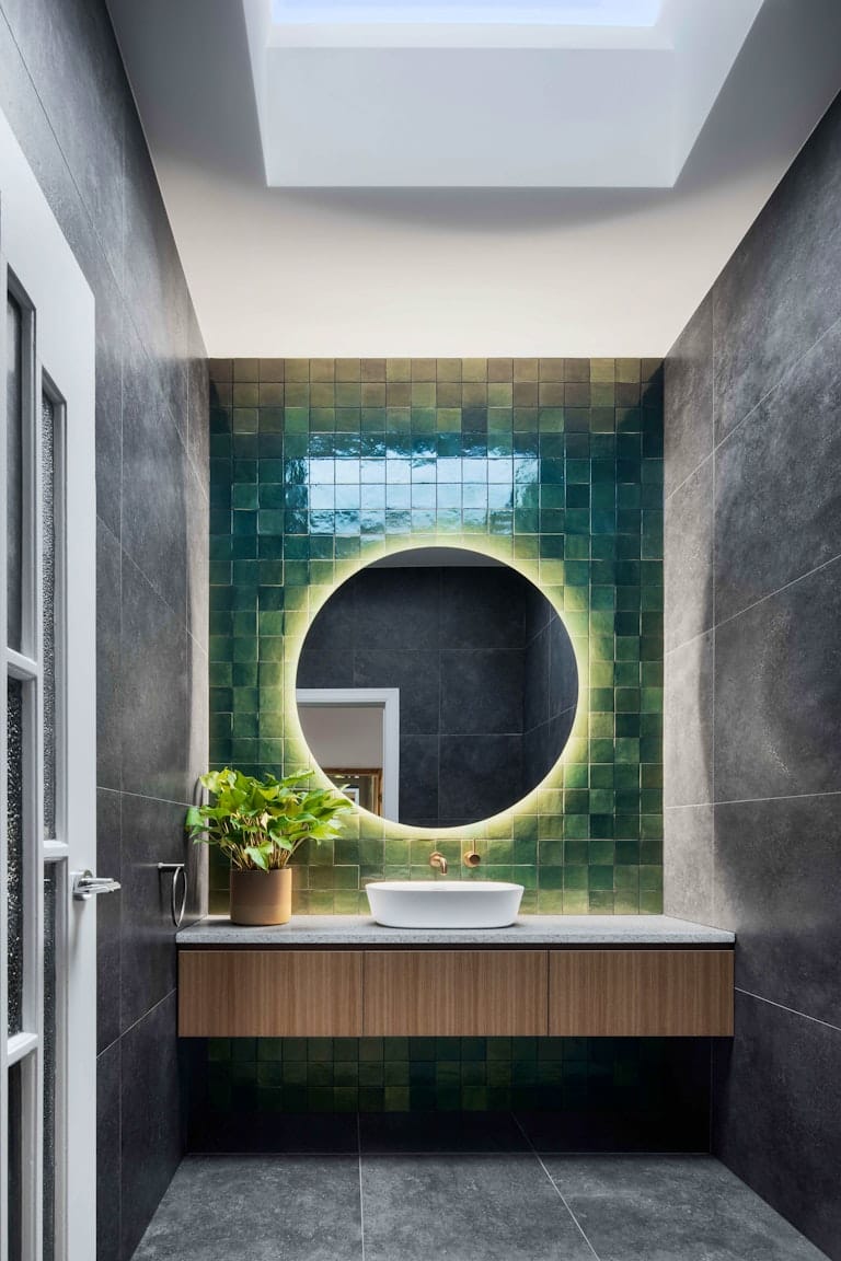 Dickens Street Residence by Chan Architecture. Photography by Tatjana Plitt. Bathroom timber vanity with dark tile floors and walls. Round wall mirror in front of green wall tiles. 