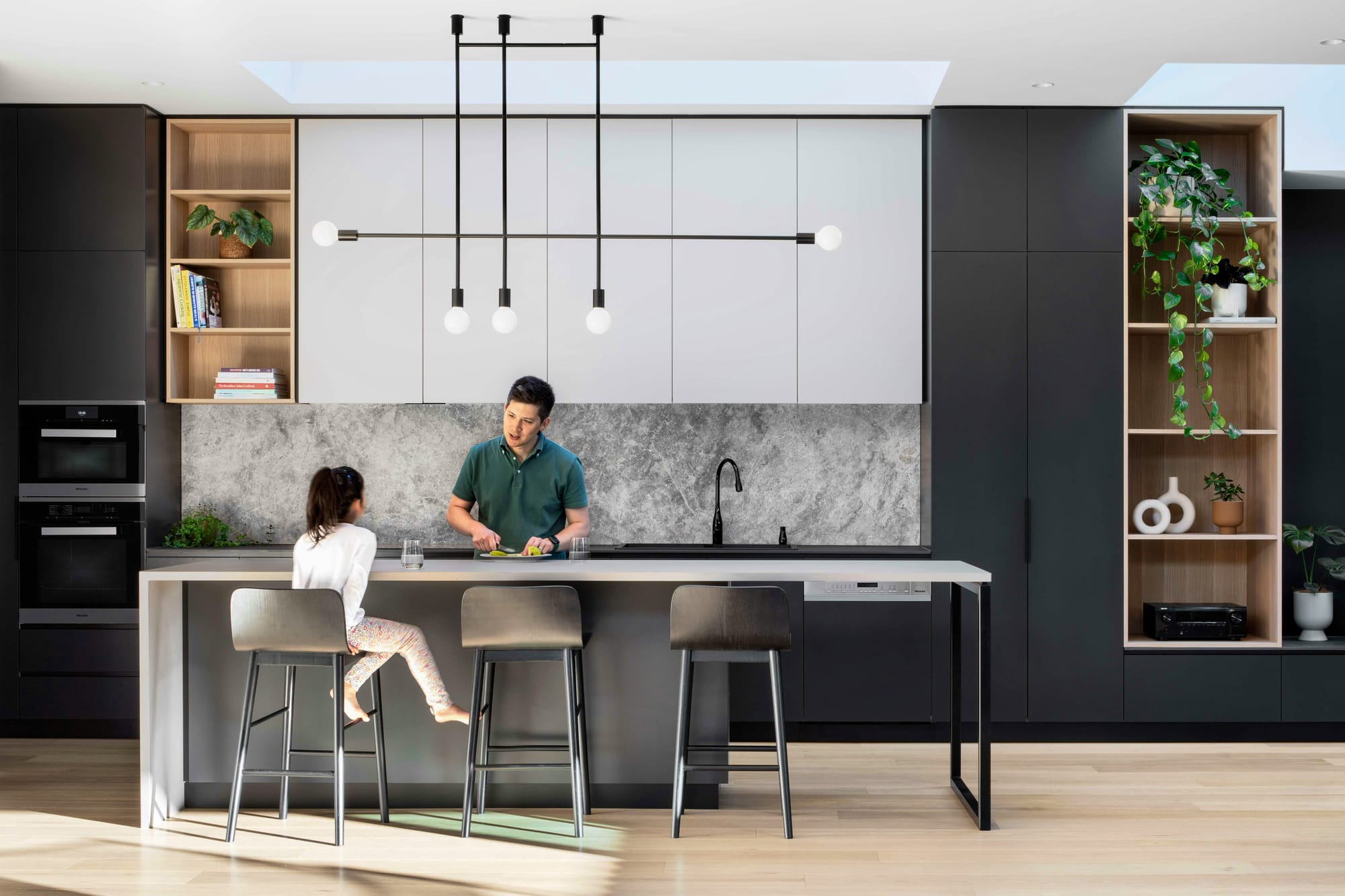 Dickens Street Residence by Chan Architecture. Photography by Tatjana Plitt. Contemporary black, white and stone kitchen with light timber floors. Young girl sitting at island bench talking to man behind counter. 