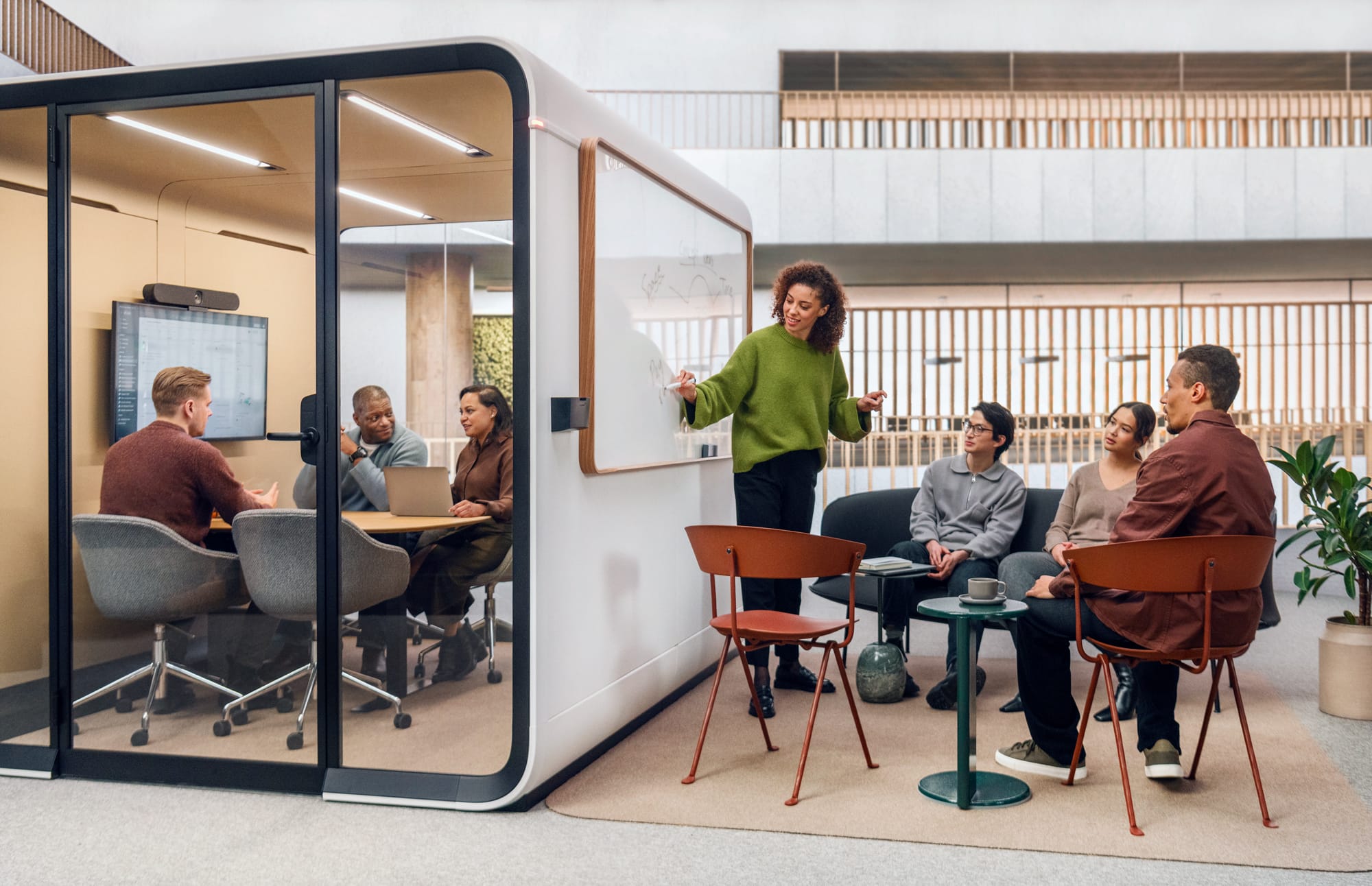 Framery Smart Pod. Copyright of Framery. Lady writing on a white board on the outside wall of a work pod. Three individuals having a meeting inside the white work pod. 