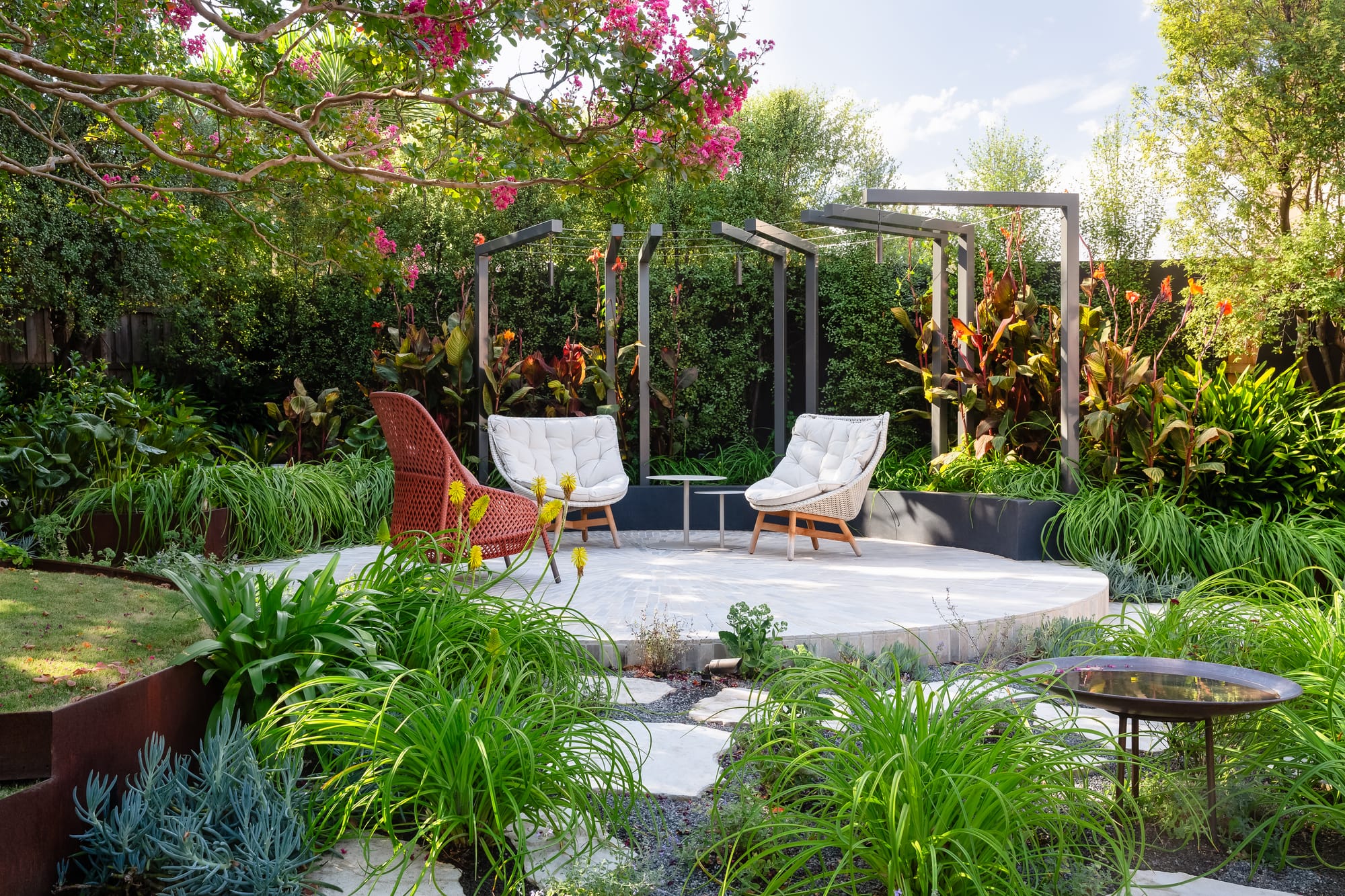 Iris Glen Iris by Julie Crowe Landscape Design. Photography by Erik Holt Photography.  Raied circular seating area in garden. Two white and one red chair underneath pergola. Surrounded by tall plants, trees and hedges.  