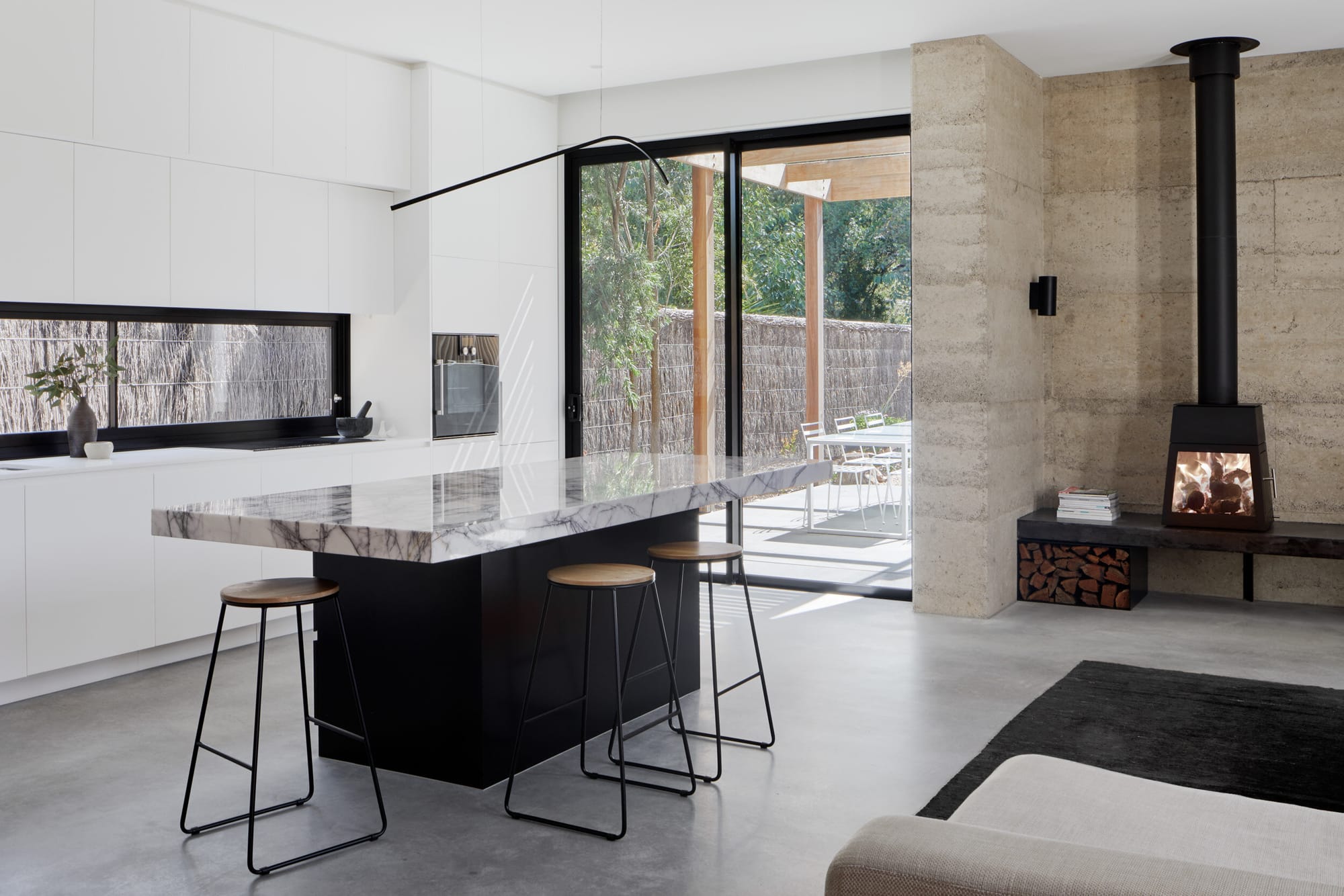 Laurel Grove by Kirsten Johnstone Architecture. Photography by Tatjana Plitt. Open plan living and kitchen space with concrete floors and rammed earth wall. Black and marble island benchtop. Black fireplace.