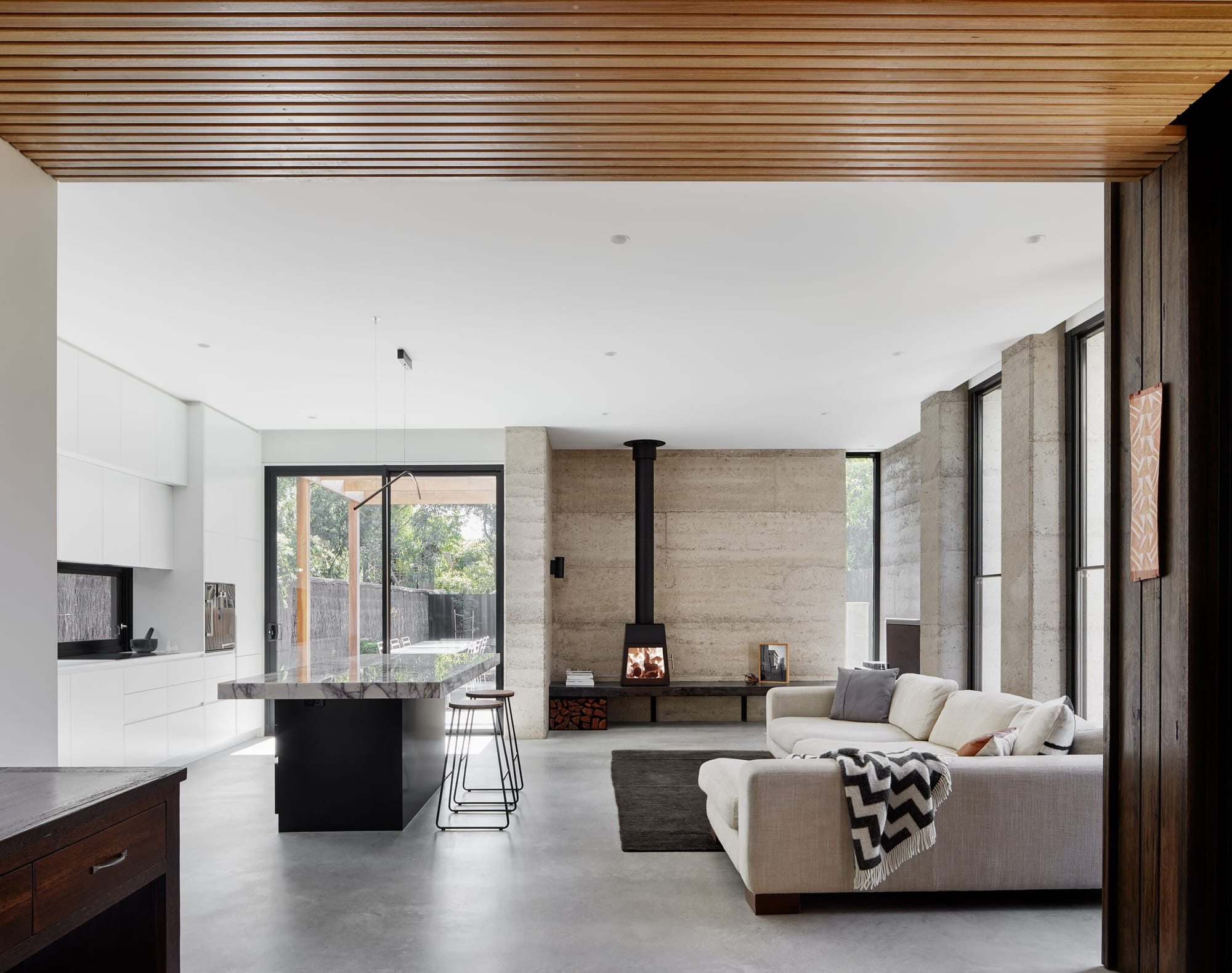Laurel Grove by Kirsten Johnstone Architecture. Photography by Tatjana Plitt. Open plan living and kitchen space with concrete floors and rammed earth feature wall behind fireplace. 