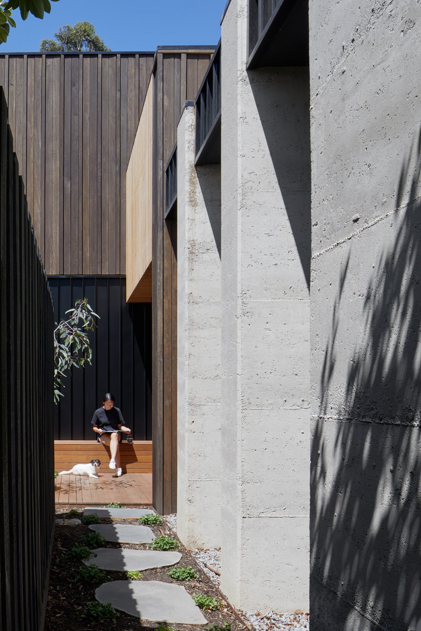 Laurel Grove by Kirsten Johnstone Architecture. Photography by Tatjana Plitt. Side of residential home with pavers leading towards timber deck and seating on left hand side. Tall concrete walls on right hand side.  