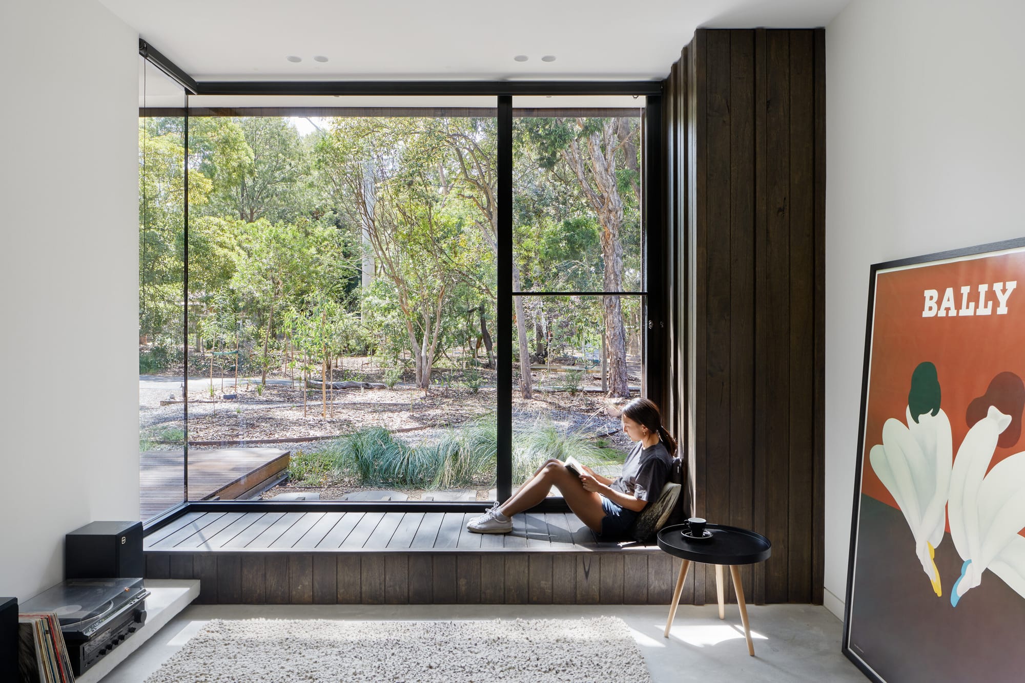 Laurel Grove by Kirsten Johnstone Architecture. Photography by Tatjana Plitt. Large window overlooks bushland surrounding house. Young girl sits on timber bench seat running underneath interior window. 
