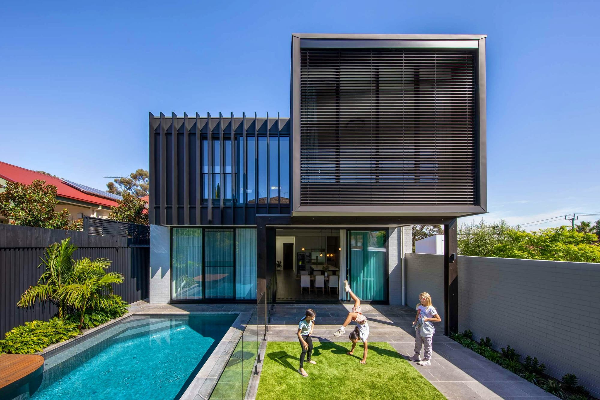 Northcote Renovation by Chan Architecture. Photography by Tatjana Plitt. Raer facade of contemporary double storey family home. Pool on left hand side of backyard. Children play on grass to right hand side of backyard. 