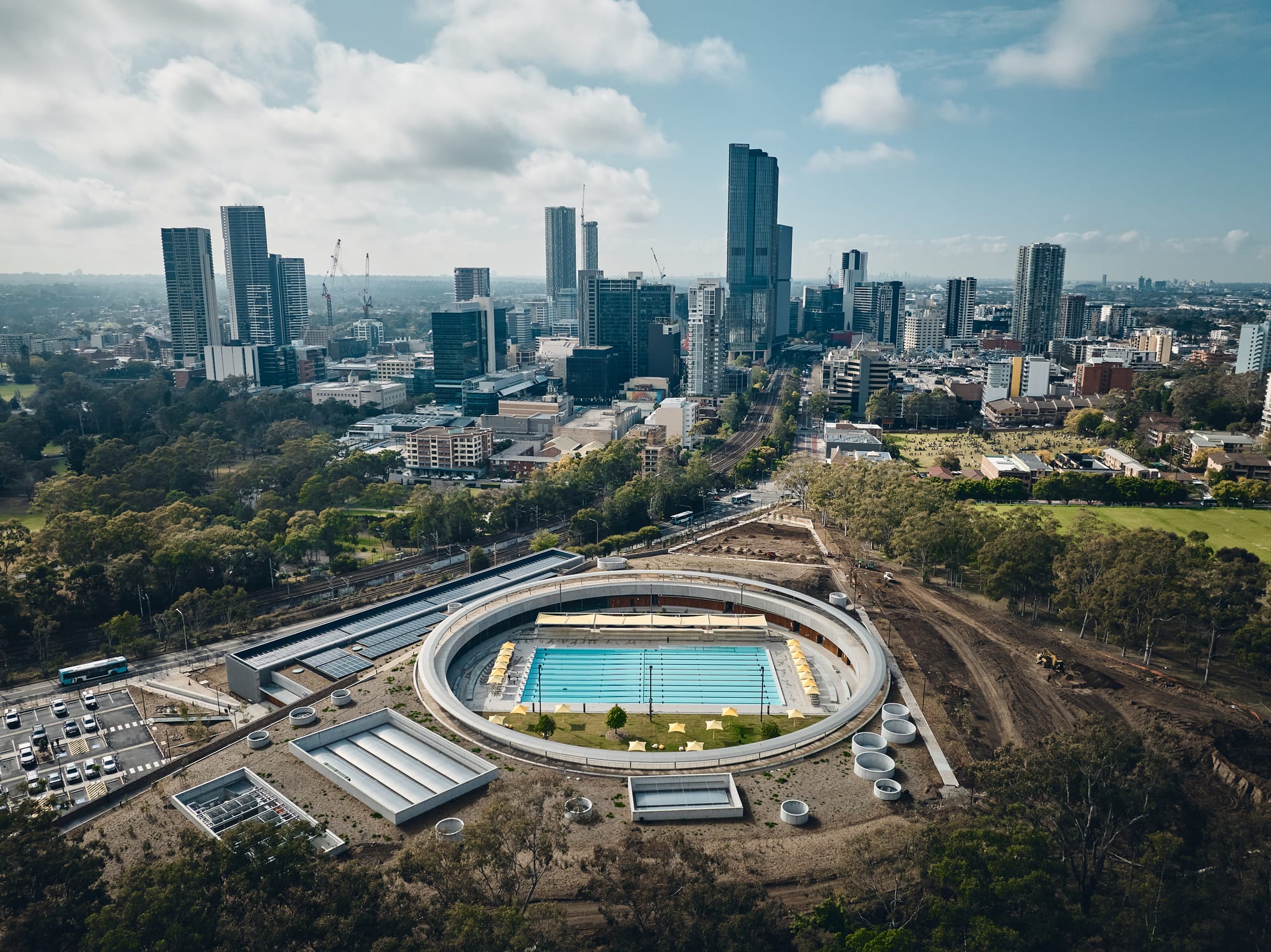 Parramatta Aquatic Centre by Mortlock Timbers. Photography by Peter Bennetts. Aerial view of outdoor public pool enclosed in large ring design. City skyline visible in background. 