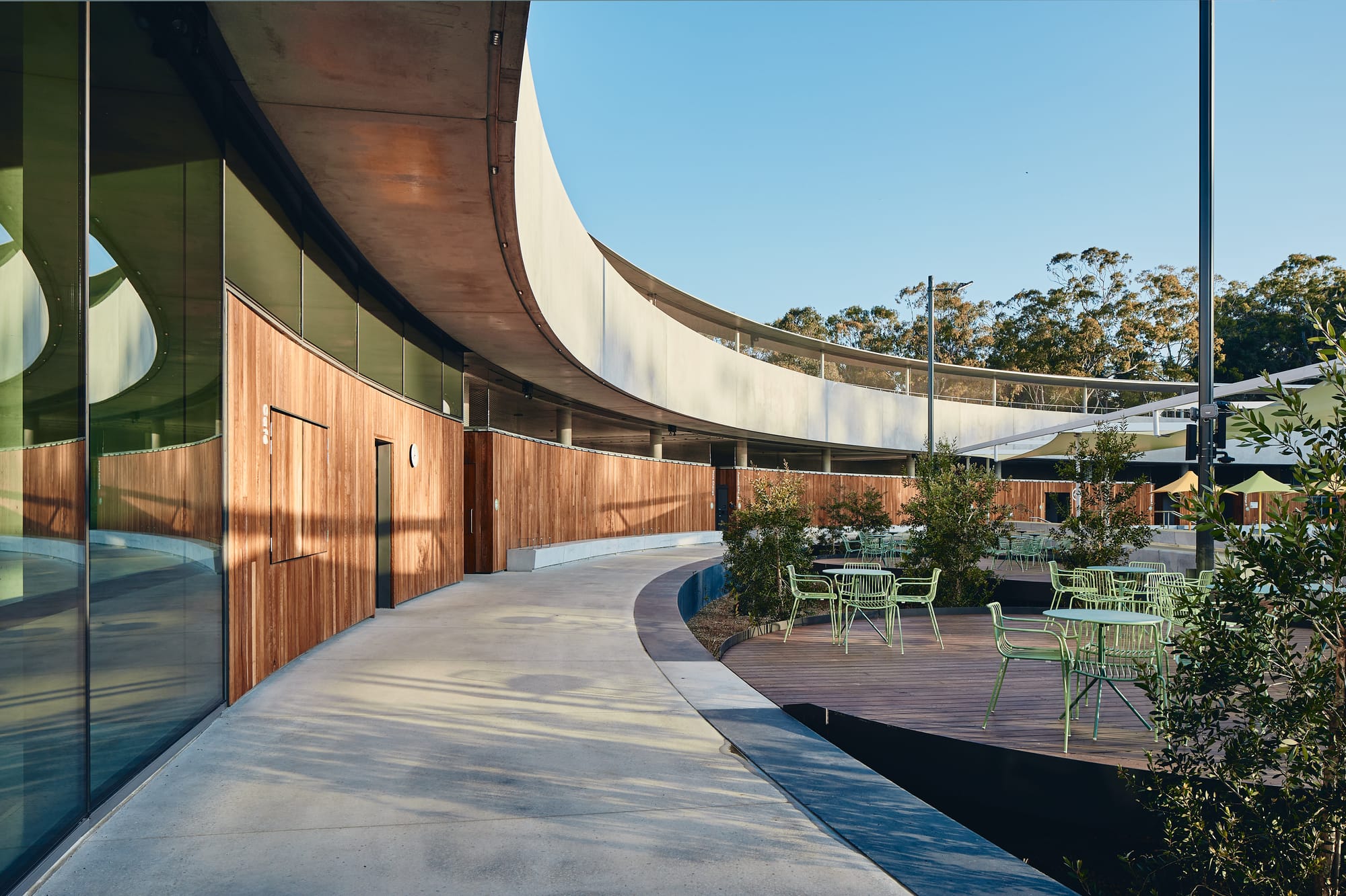 Parramatta Aquatic Centre by Mortlock Timbers. Photography by Peter Bennetts. Curved outdoor walkway clad in timber. Timber deck deating area. 