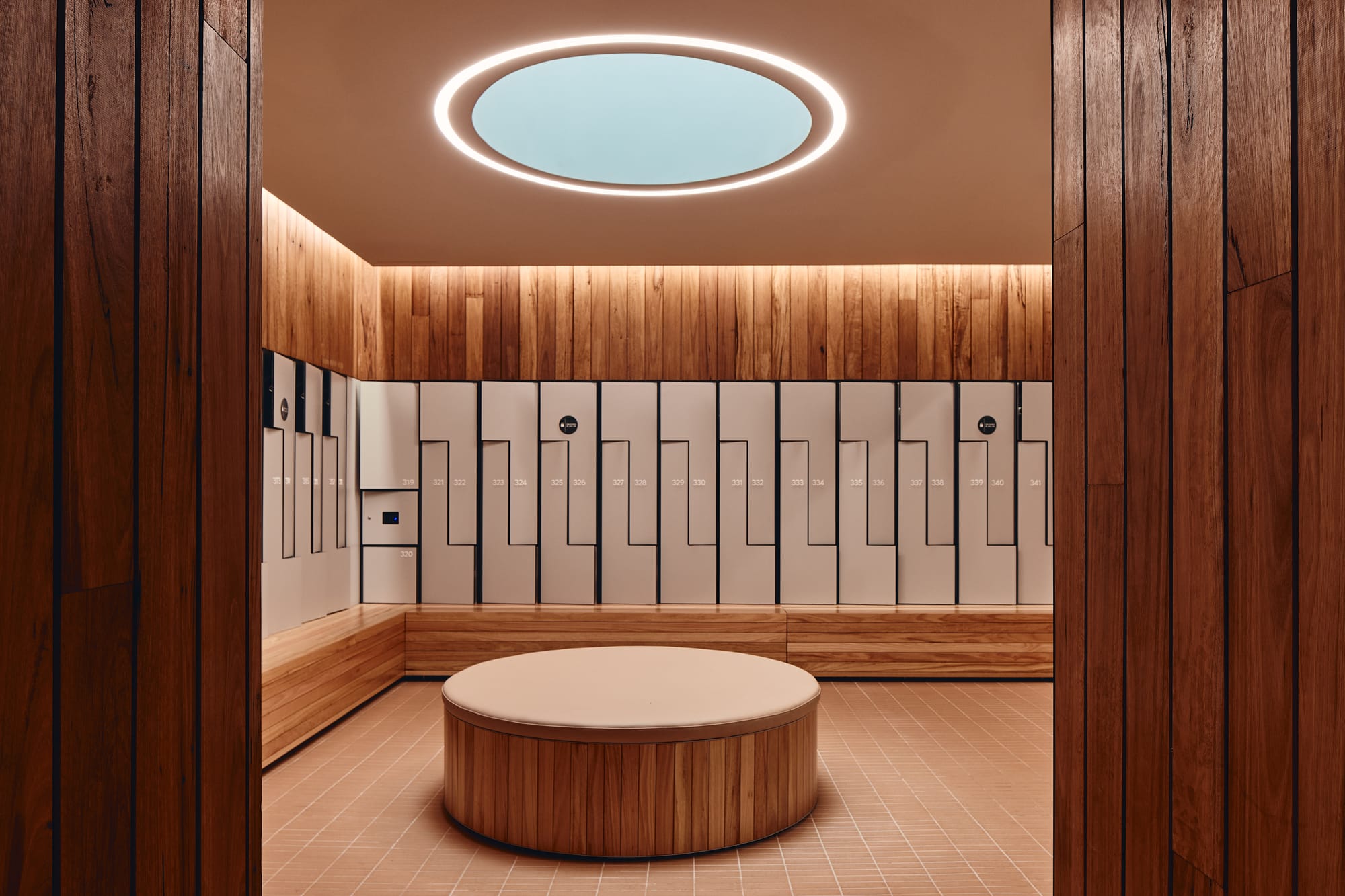 Parramatta Aquatic Centre by Mortlock Timbers. Photography by Peter Bennetts. Indoor locker room with round timber seat in centre of room. Timber clad walls, tile floors and white lockers. 