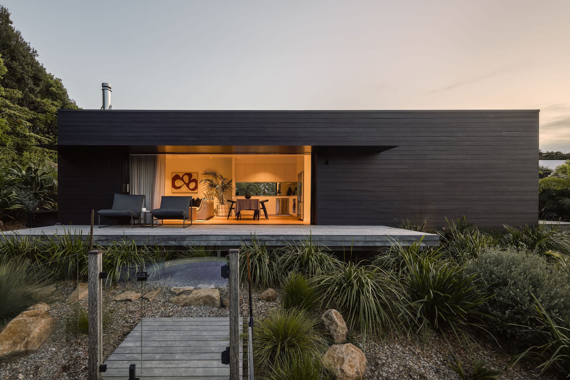 Rockpool Farm Byron Bay. Photography by Andy Macpherson. Rectangular black timber clad building with extending aged-timber deck. Large opening from deck to dining and living space. Warm internal lighting.