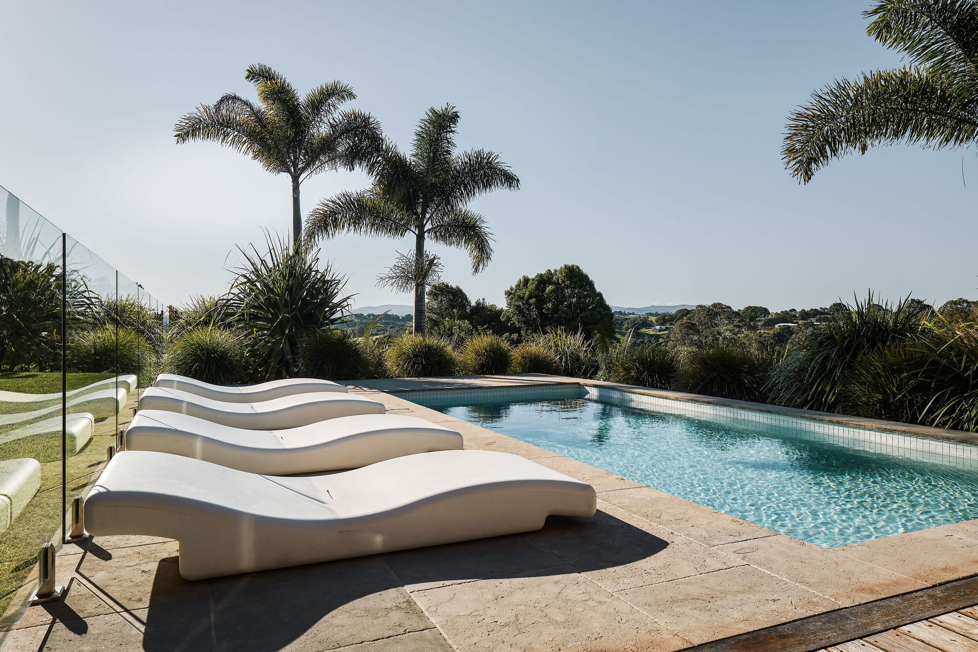 Rockpool Farm Byron Bay. Photography by Andy Macpherson. White chaise lounge recliners by outdoor pool. Stone pavers. Views of hills and plants behind pool.