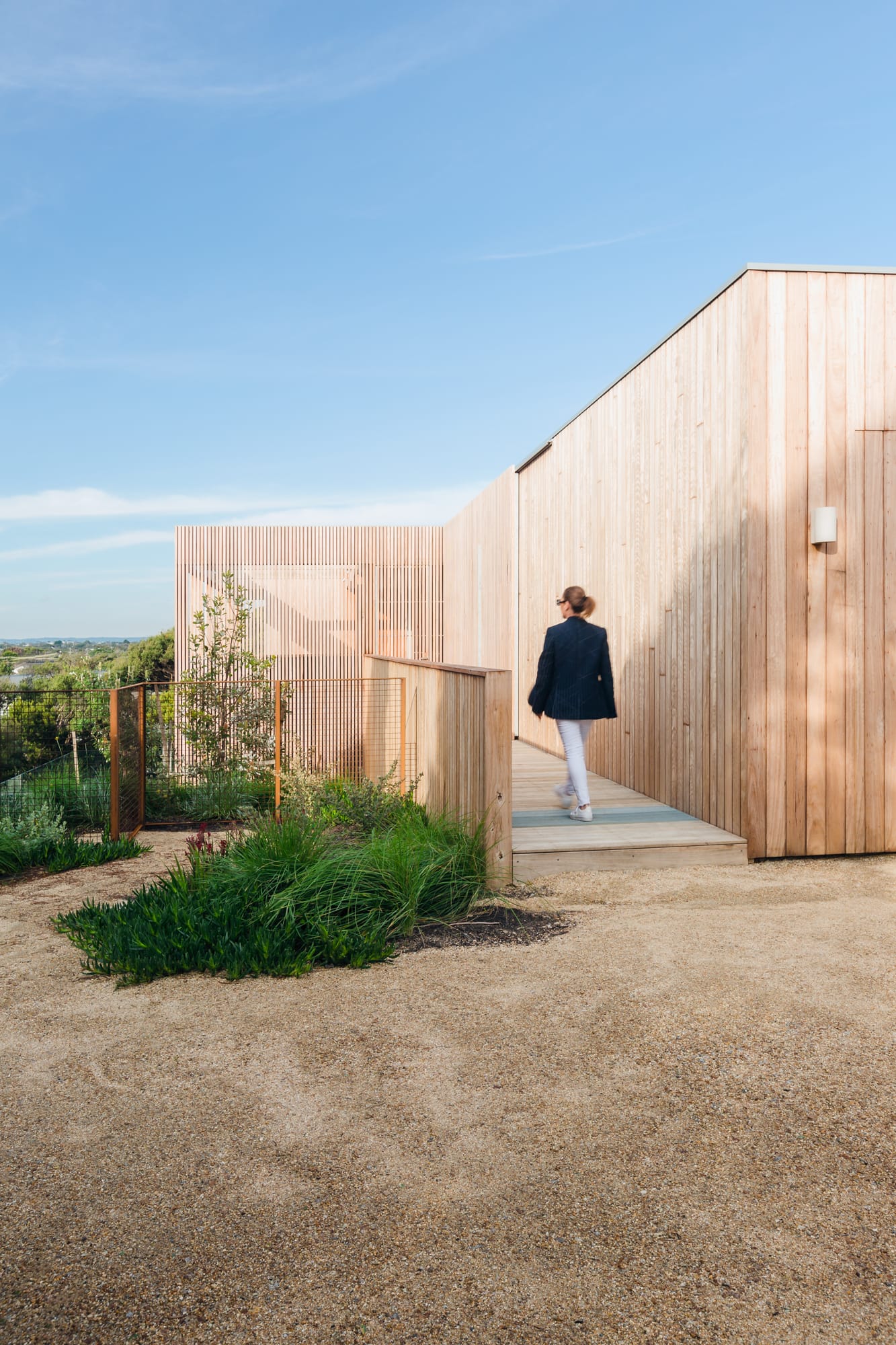 Ritchie Blairgowrie by Julie Crowe Landscape Design. Photography by Erik Holt Photography.  Women walks across timber deck running alongside timber-clad home. Crushed limestone flooring below deck. Native plant life. 