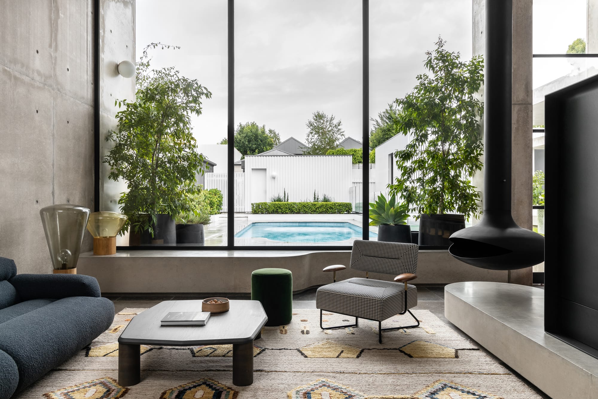 Rose Park by studio gram. Photography by Timothy Ross. Living room with Aztec print rug and floor-to-ceiling black framed windows overlooking garden and pool. Black dome fireplace hanging from ceiling. Concrete bench running along window. 