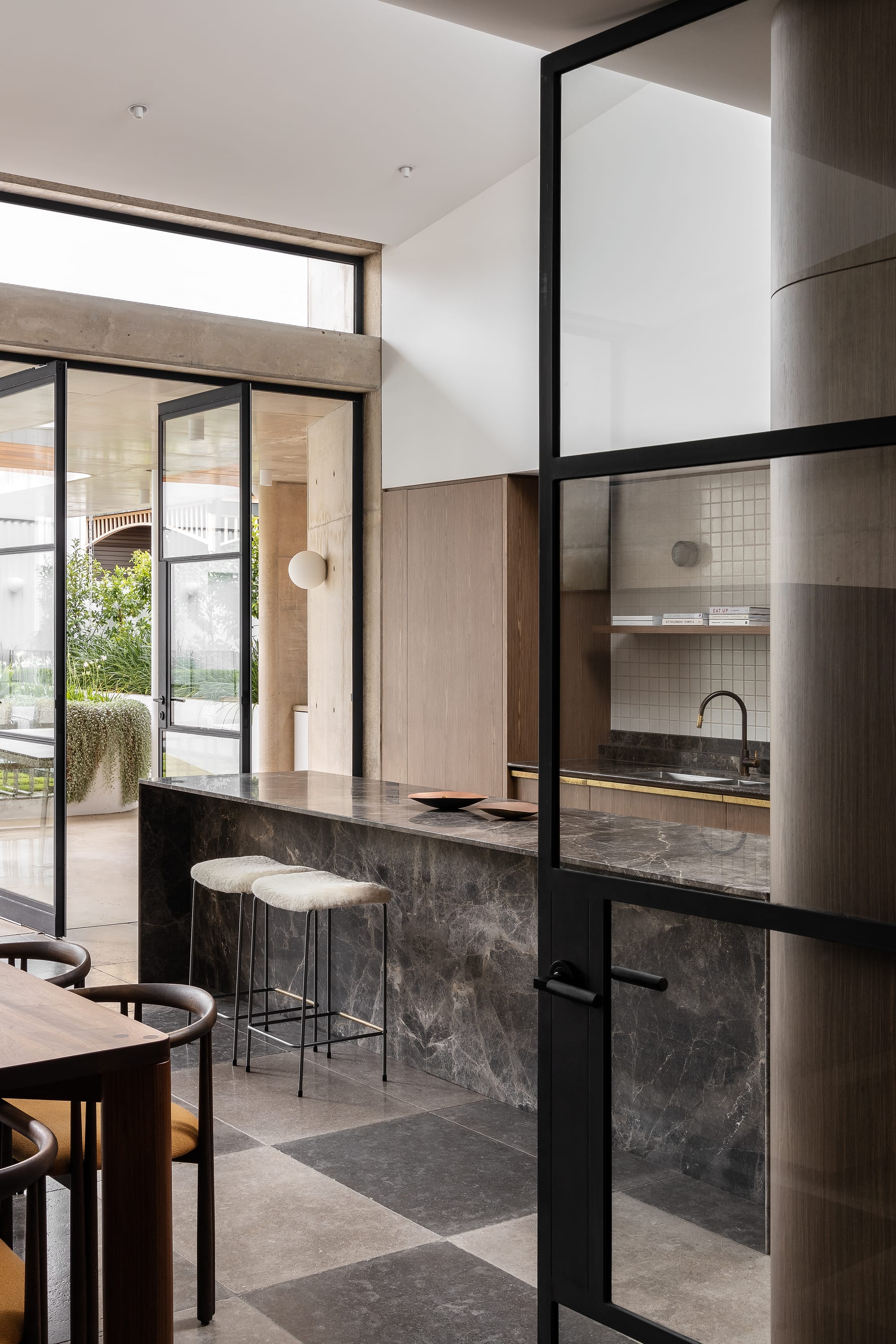 Rose Park by studio gram. Photography by Timothy Ross. Dark grey stone counters and countertops in kitchen. Black framed glass doors open onto undercover patio. Checkered floor tiles. 