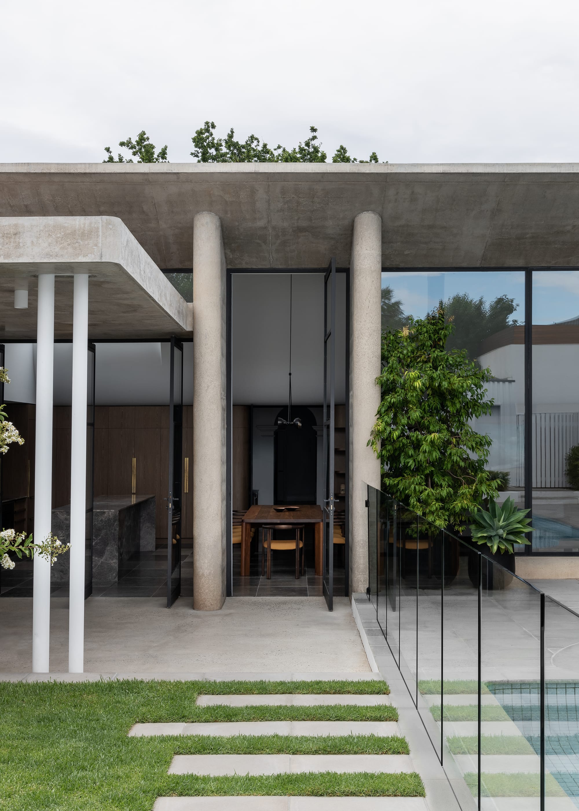Rose Park by studio gram. Photography by Timothy Ross. Rear facade of concrete home. Steps leading from back verandah across grass. Floor to ceiling glass windows and doors. 