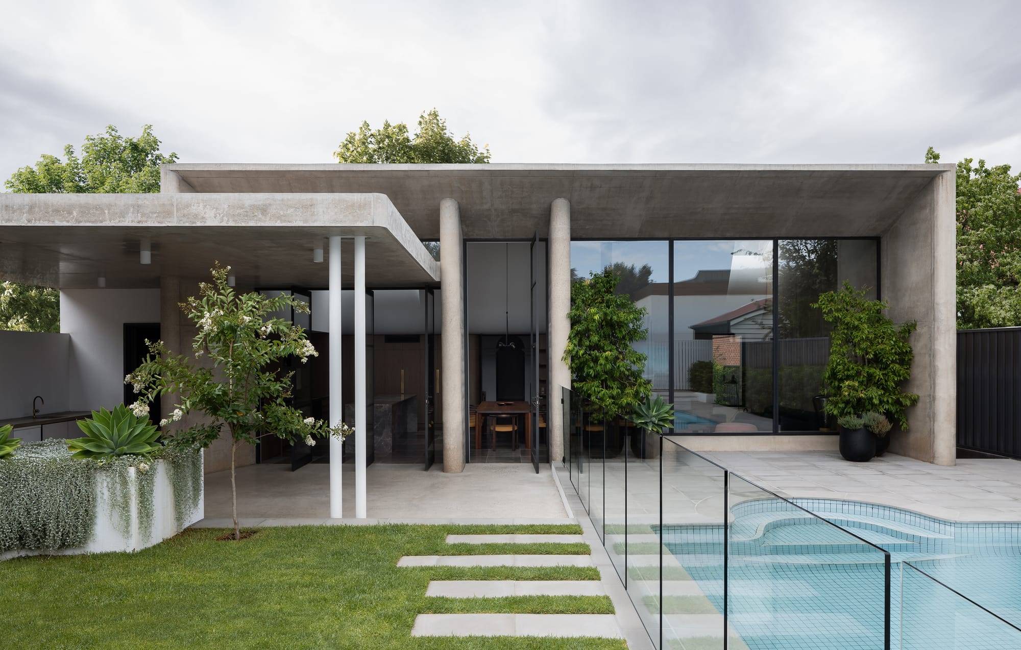 Rose Park by studio gram. Photography by Timothy Ross. Rear facade of residential home constructed of concrete. Green grass with pavers. Tiled pool with glass fence to right. 
