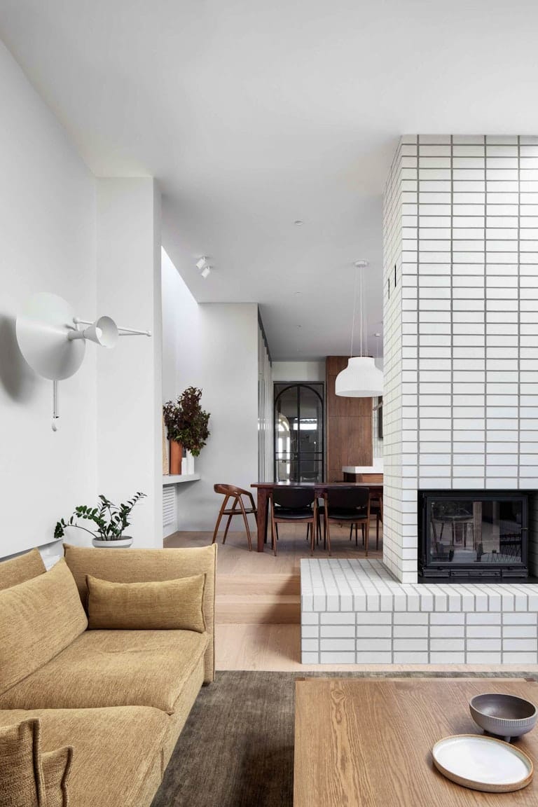 South Terrace Renovation by Chan Architecture. Photography by Tatjana Plitt. Sunken living room with white brick fireplace and muted yellow couch. Small step leading up to dining area in background. 