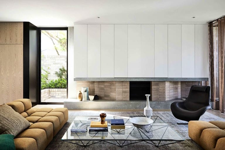 The Split Home by Seidler Group. Copyright of Seidler Group. Living room in residential home with white, brick and concrete fireplace running length of wall. Glass and metal coffee table in front of mustard couch and black armchair. 