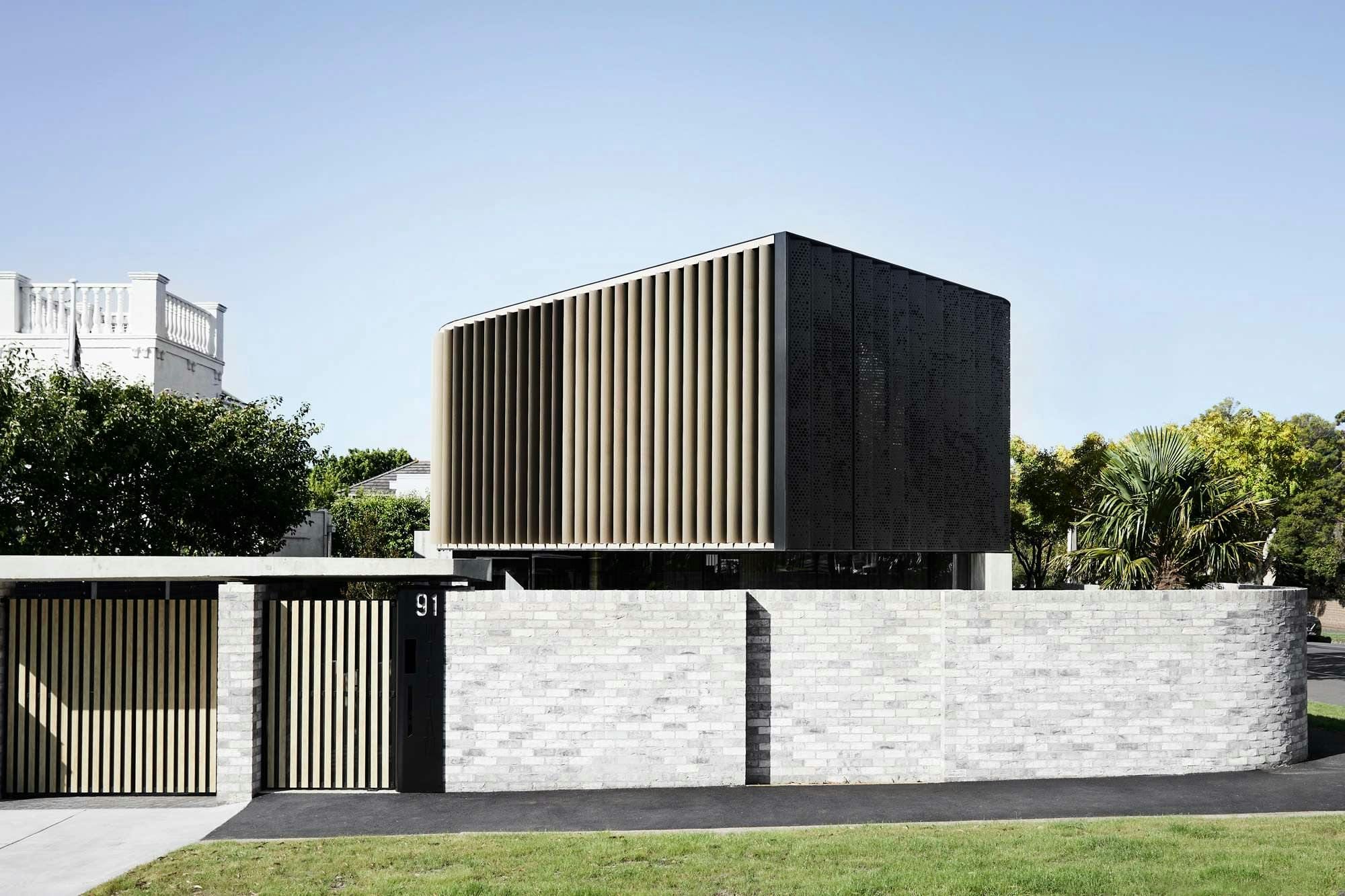 The Split Home by Seidler Group. Copyright of Seidler Group. Street view of homes facade. Brick fence surrounds property with timber gate in left hand corner. Second storey of home wrapped in black metal and visible above fence. 
